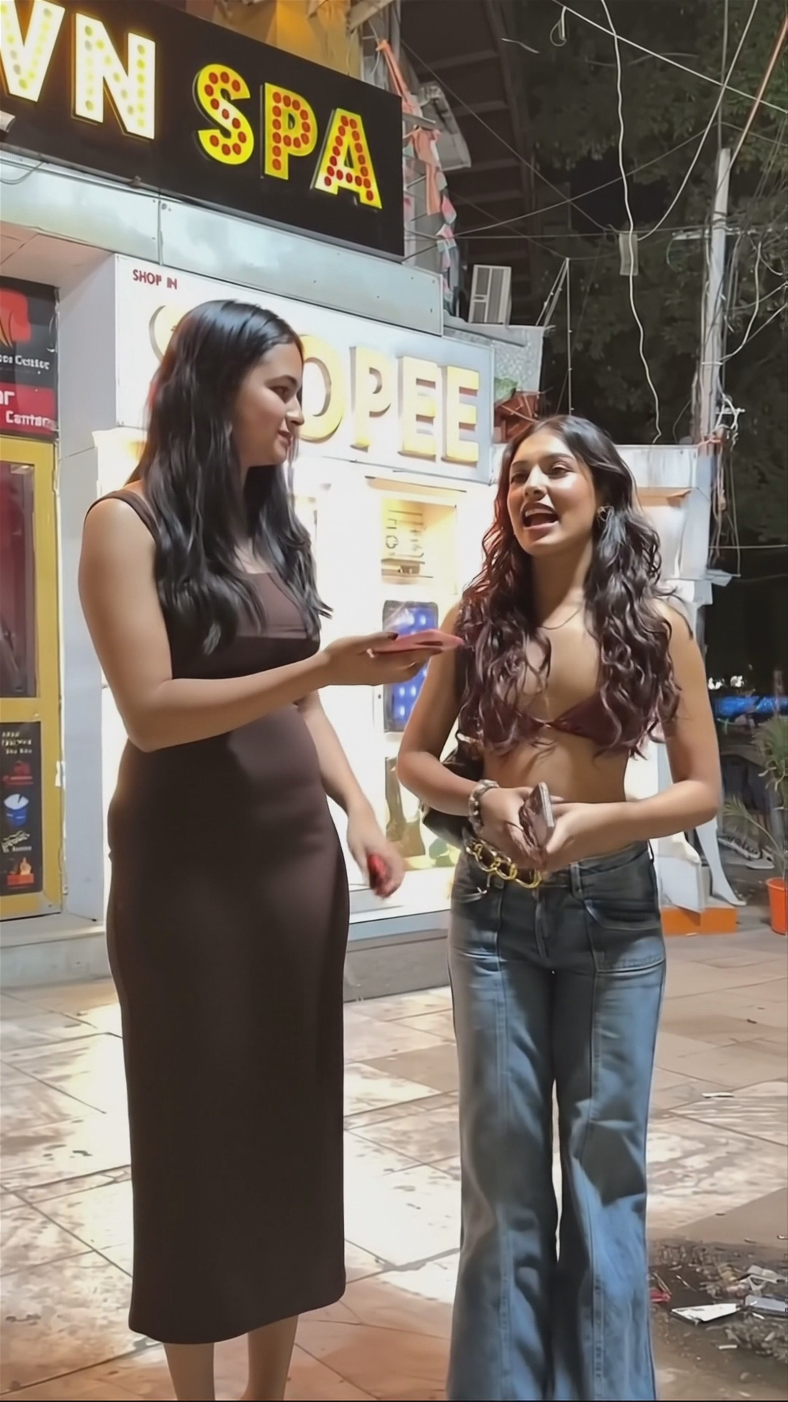 A vertical 1080x1920 video still generated by Veo 3 shows two young women during a street interview at night. The woman on the left wearing a brown dress holds a microphone, while the woman on the right in a crop top and jeans listens. Bright spa signage glows in the background.