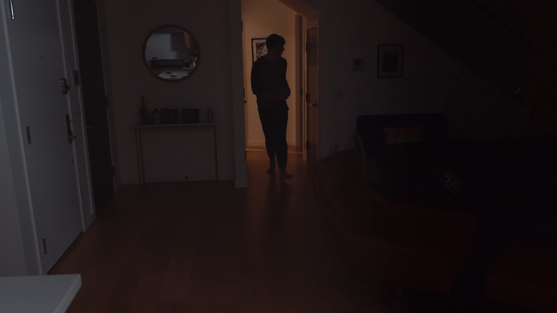 A man struggles to sleep in his bedroom before getting out of bed and walking into a hallway to check his smartphone. His movements reflect restlessness and the quiet atmosphere of a late night at home.