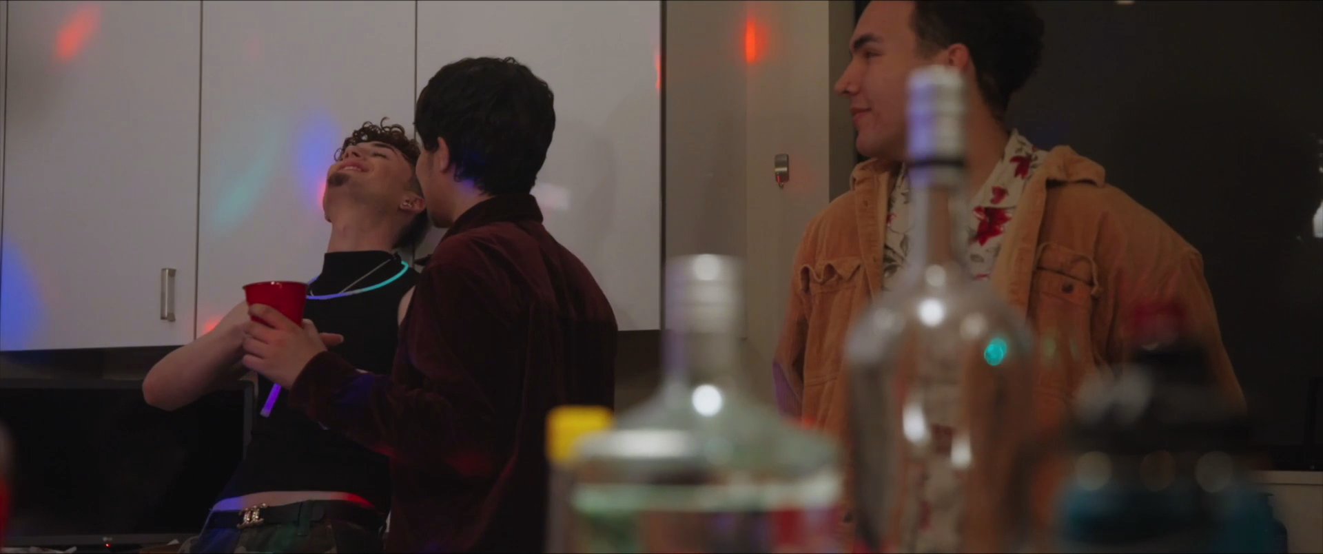 A group of young adults socialize and dance in a kitchen setting during a house party atmosphere. The scene captures a lively, casual evening interaction marked by festive lighting and shared drinks.