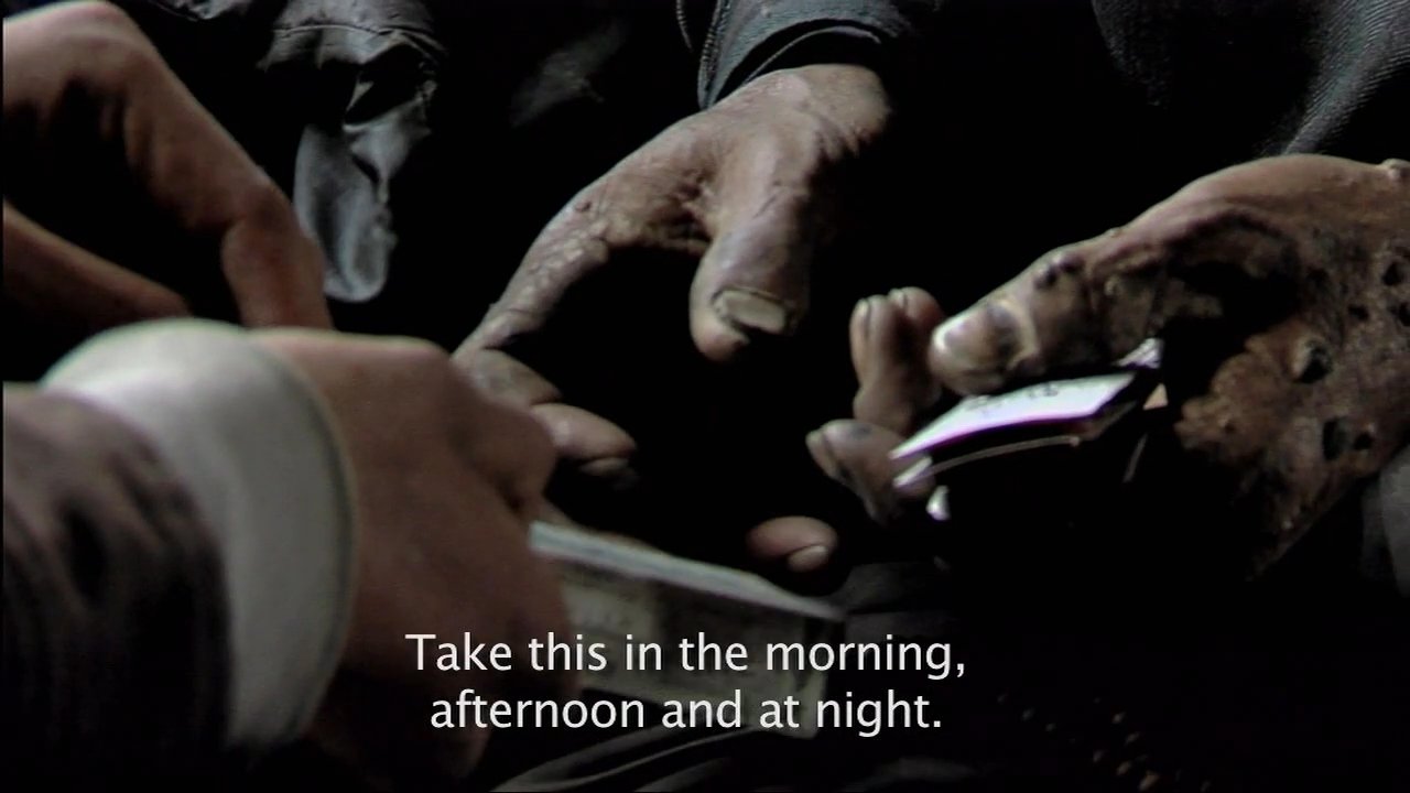 A healthcare professional performs a medical examination on an elderly patient in a remote, dimly lit dwelling, providing instructions for their medication. The scene transitions to the surrounding arid landscape, highlighting the isolation of the rural medical setting.