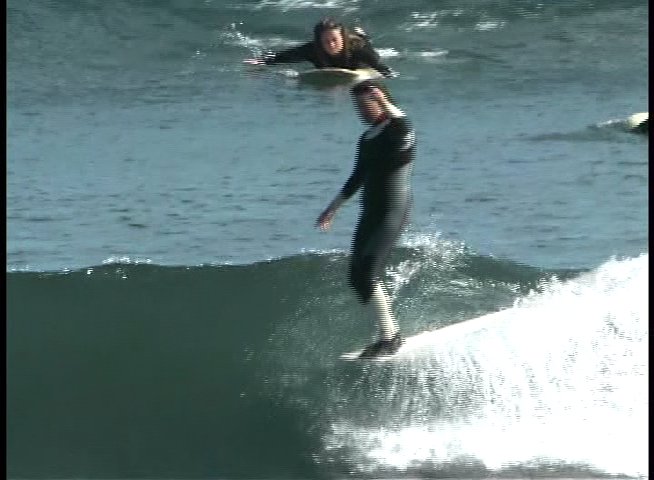 Surfers navigate rolling ocean waves in a coastal setting while wearing wetsuits. The scene captures various individuals riding the surf against a backdrop of distant marine vessels.