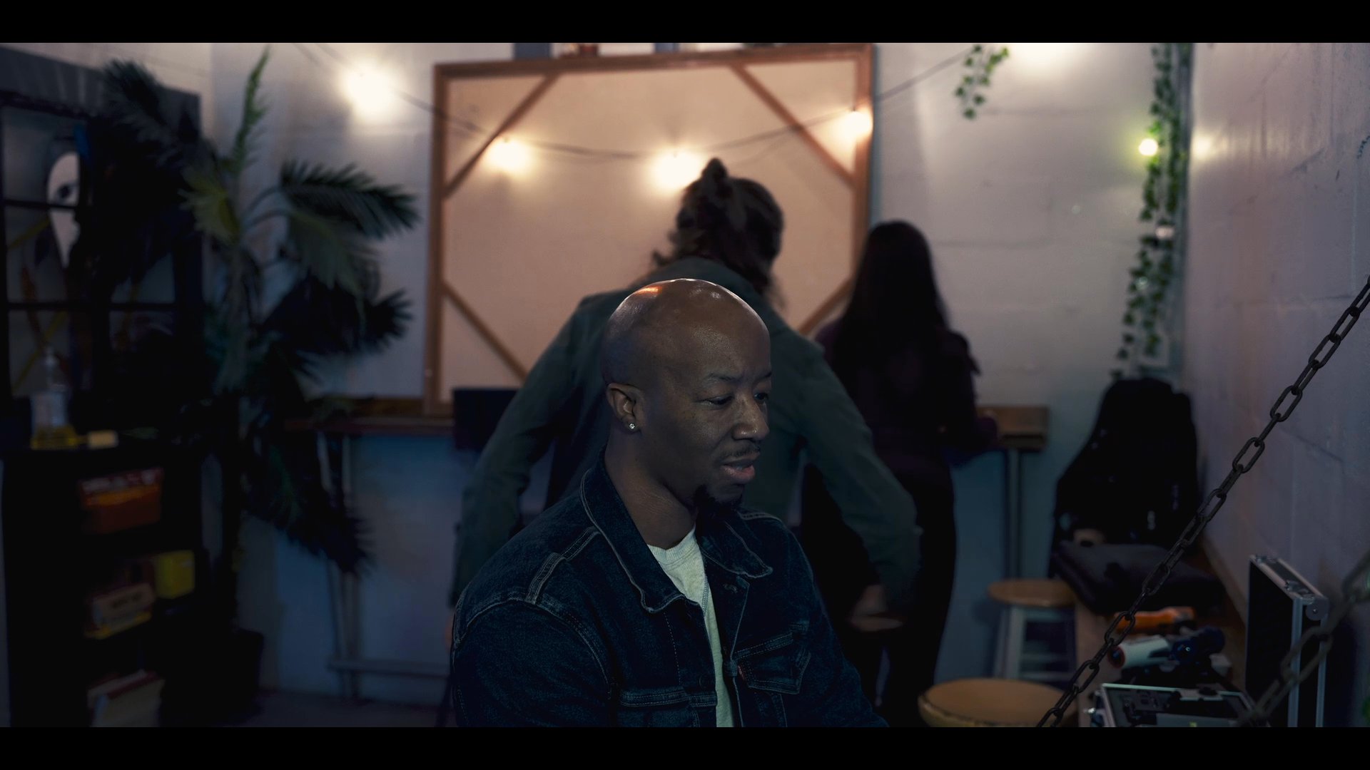 Three individuals engage in a brief discussion within a dimly lit room before one person transitions to resting on a nearby sofa.