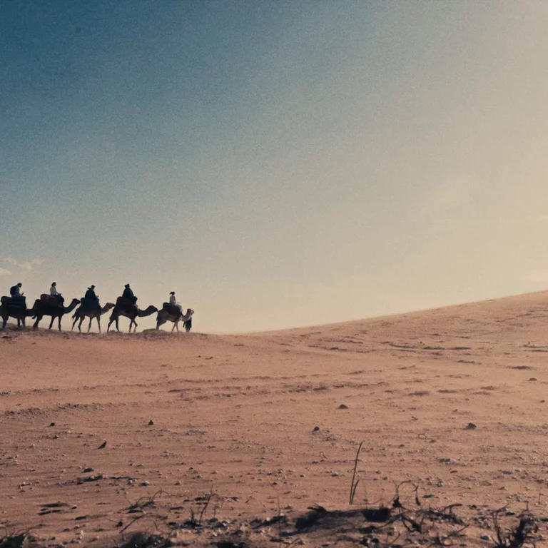 Video of caravan of people riding camels into the distance of a wind-blown Moroccan desert