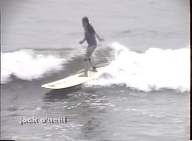 Historical footage captures a bustling surf shop exterior followed by a surfer catching waves in the ocean, honoring the legacy of Jack O'Neill. The sequence concludes with vintage street views, providing a glimpse into the early California surfing lifestyle.