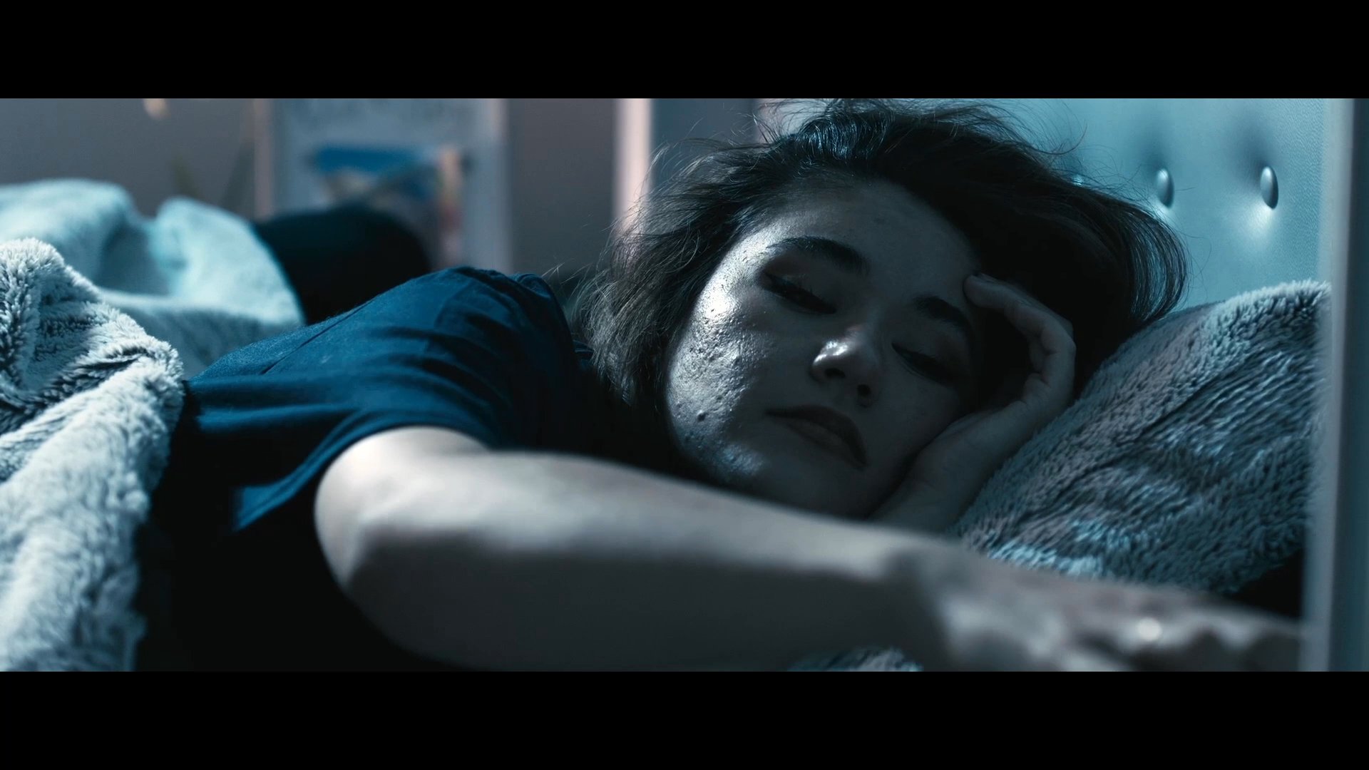 A woman struggles with restless sleep in a dimly lit bedroom as she tosses and turns under covers. She shifts positions repeatedly, unable to find comfort throughout the night.