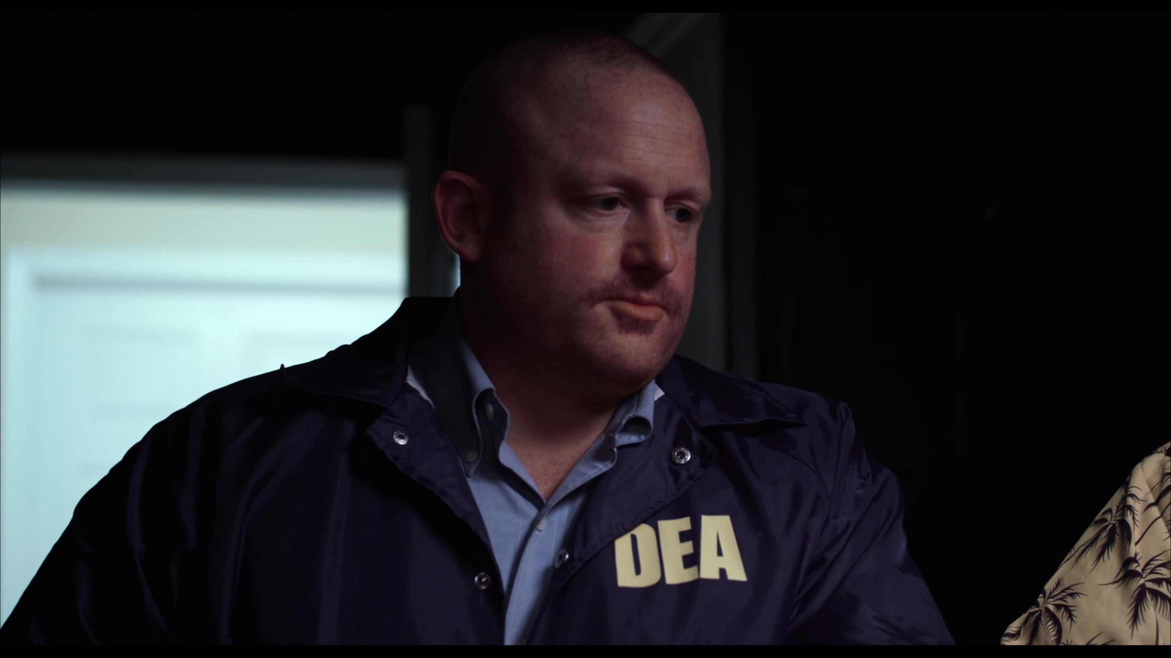 A DEA agent questions individuals in custody, facilitating a tense exchange between law enforcement and suspects in a dark, confined setting.
