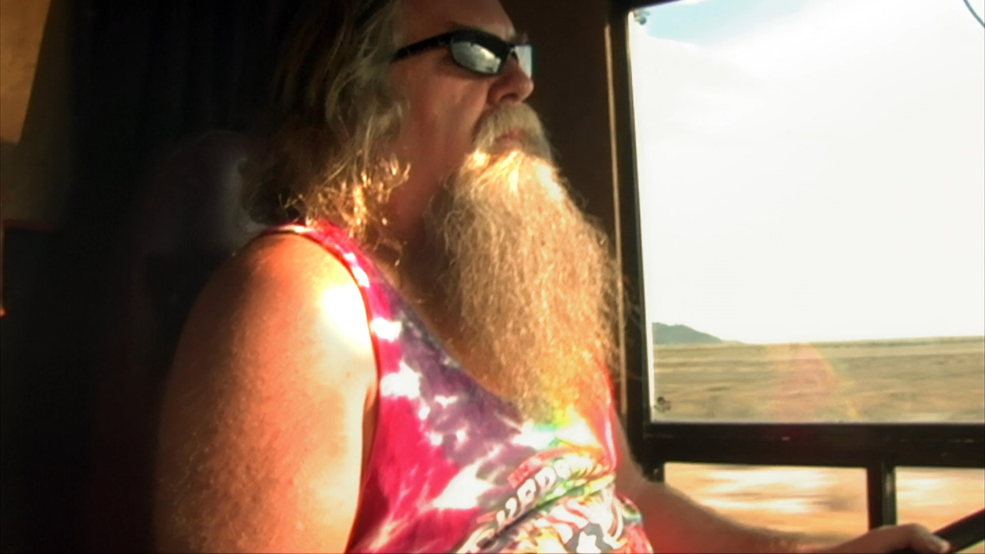 Sonny Barger, founder of the Oakland Hells Angels, discusses motorcycle culture while seated on his bike in a desert landscape, alternating with a bearded man observing the scenery from inside a vehicle.