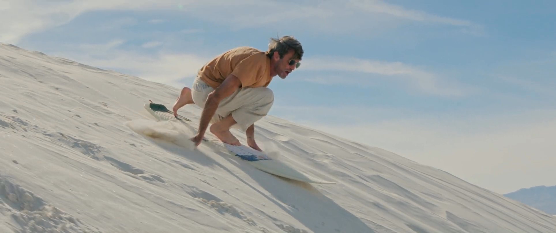 A man navigates a steep white sand dune on a board, losing his balance and tumbling before regaining control to continue the descent. The clip highlights the physical challenge and dynamic motion inherent in sandboarding.
