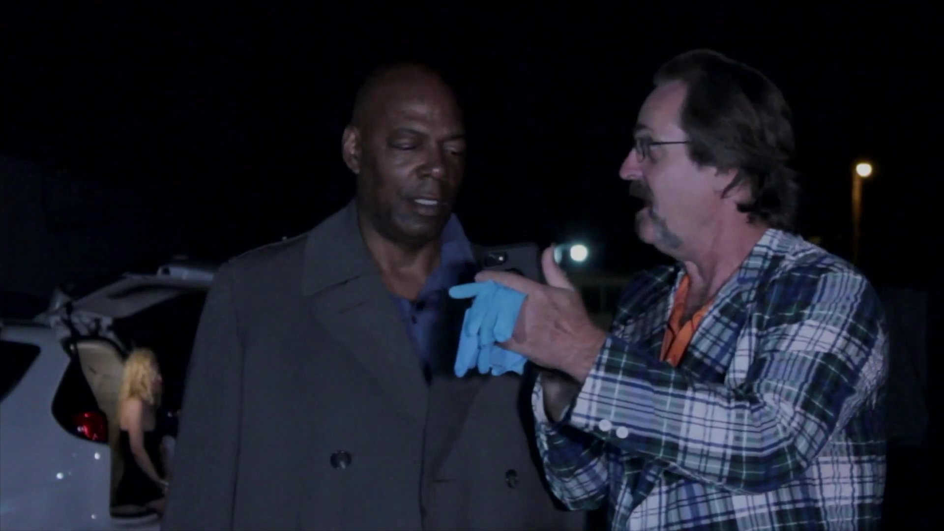 Two detectives examine digital evidence on a smartphone at a nighttime crime scene cordoned off with police tape. They discuss findings while reviewing photographs of individuals on the device.
