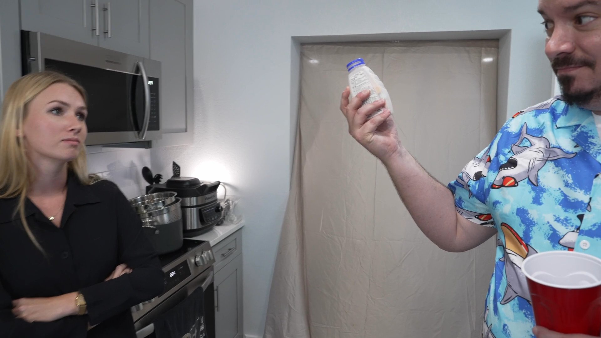 Individuals engage in a conversation within a home kitchen setting while holding drinks and navigating domestic social dynamics.