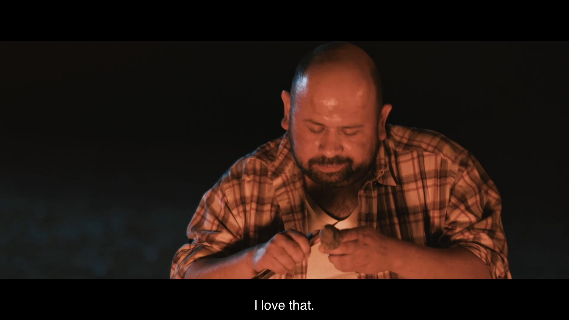 Two men converse beside a campfire at night while one of them carves wood, reflecting on the value of their creative pursuits versus financial concerns.