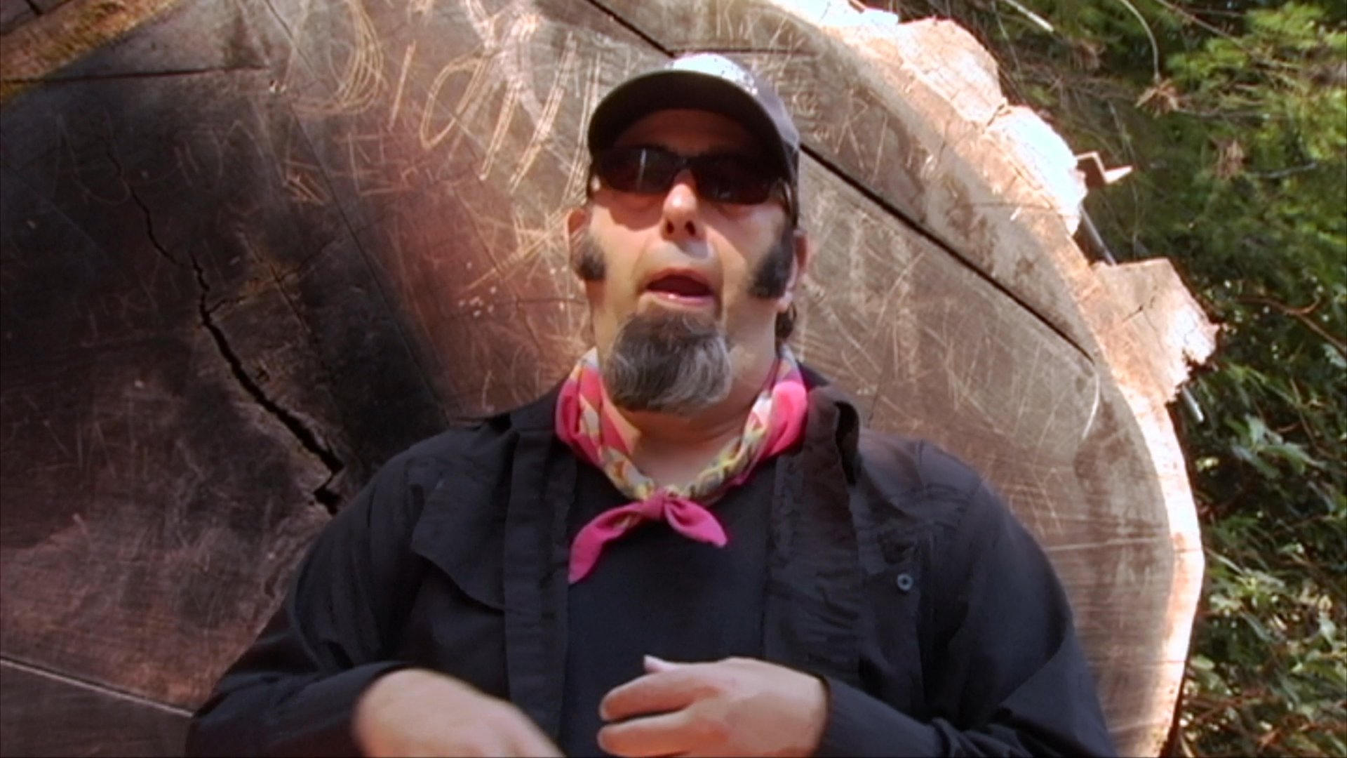 A man wearing sunglasses and a bandana speaks to an off-camera interviewer while standing in front of a large, weathered log in a forest environment.