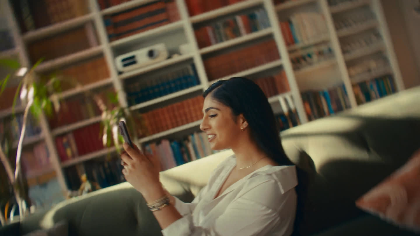 Film still: a woman smiling at her phone while sitting on a sofa