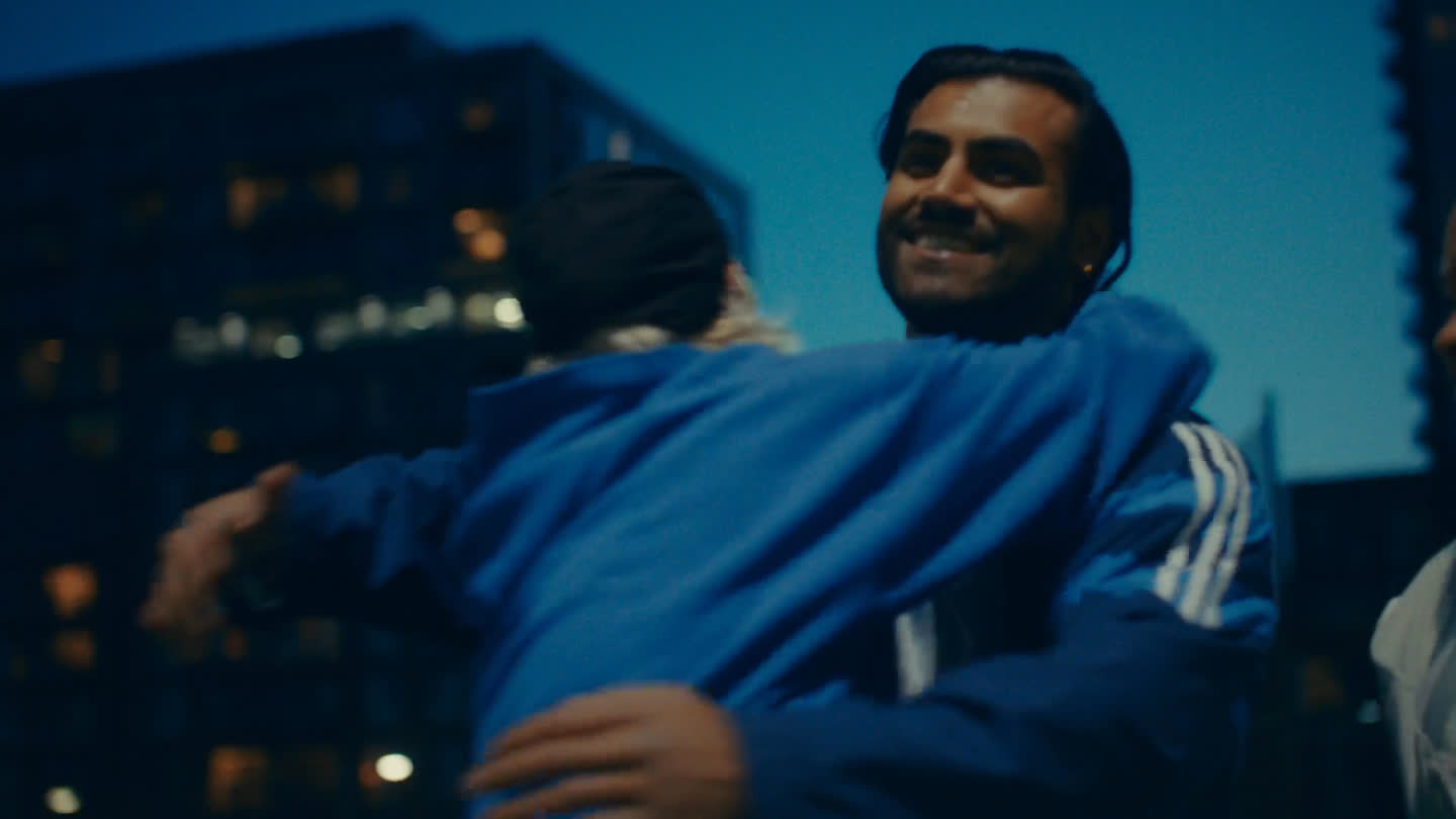 Film still: a smiling man greeting a friend with a hug outside at dusk