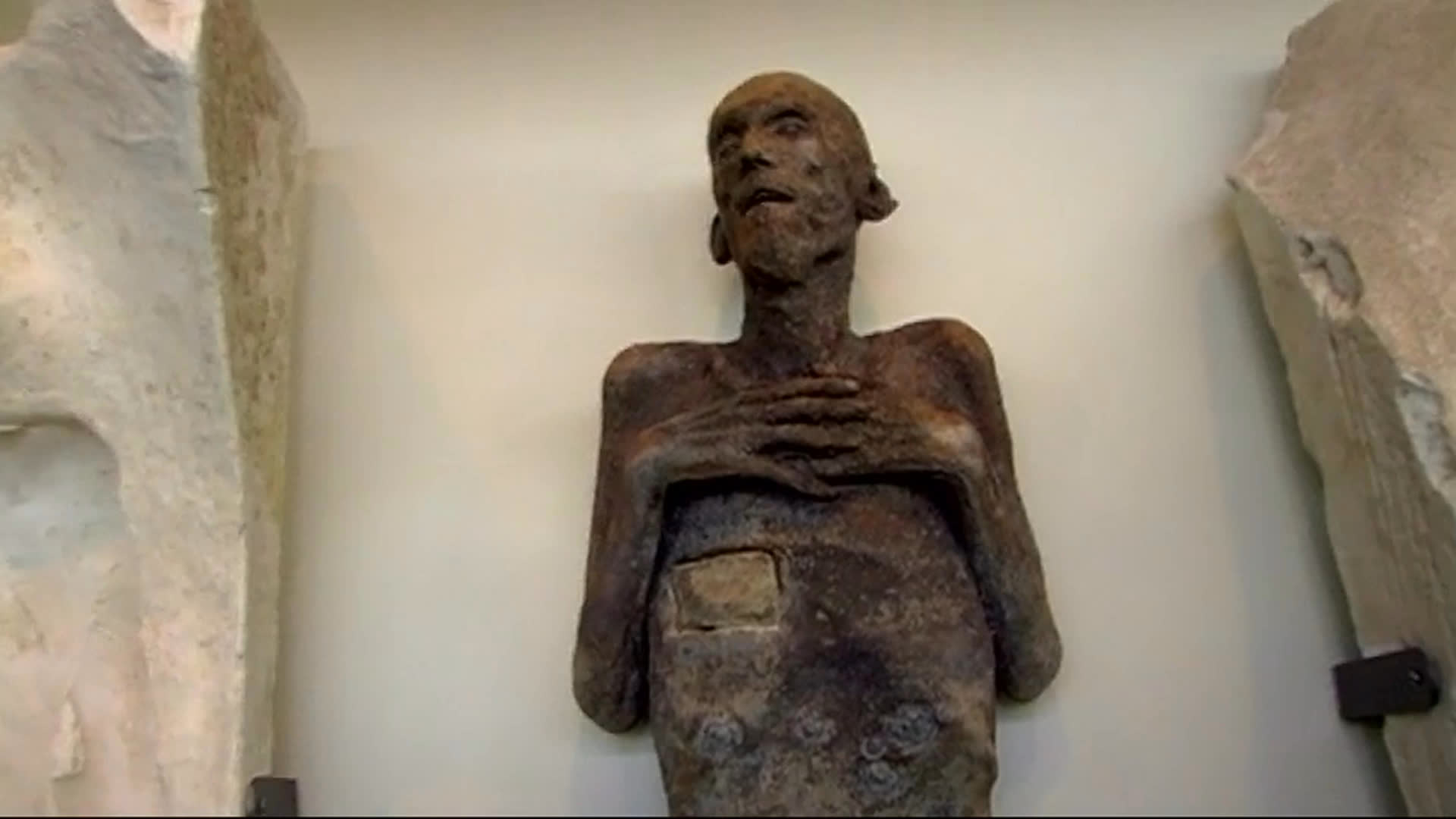 An elderly man in a green cap guides a visitor through a gallery, describing a dark human-form sculpture made from his deceased friend’s body and noting that the friend’s ashes remain inside. The tour also features a pale sculpted figure mounted on stone.