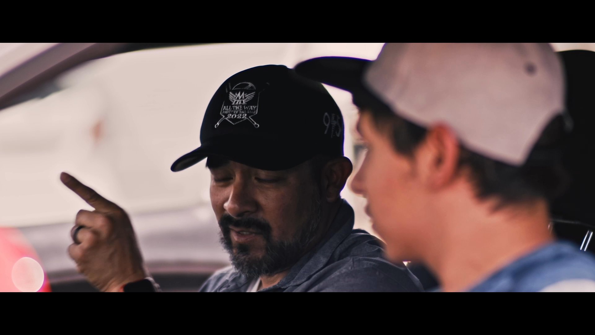 An experienced driver provides guidance and instruction to a younger student while sitting inside a sports car. The mentor gestures and speaks while the student focuses on the road, concluding with the student preparing to drive.