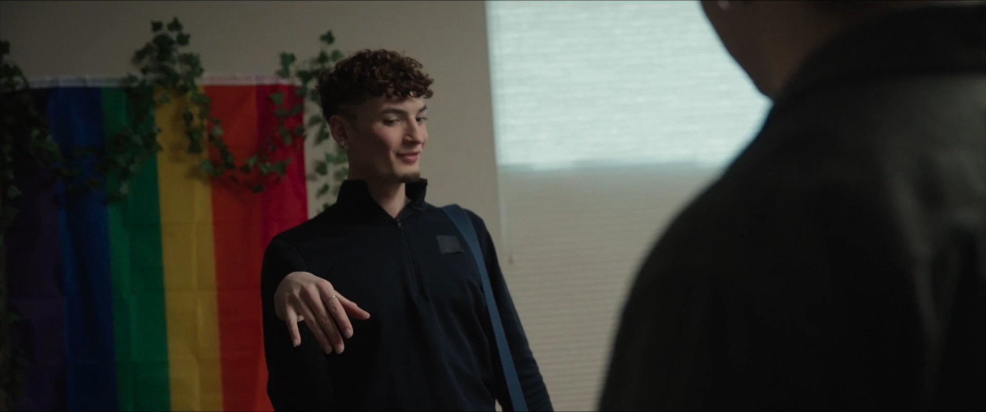 Two friends greet each other with an embrace in a bedroom before engaging in an animated conversation. The setting includes a pride flag, highlighting a personal and welcoming atmosphere.