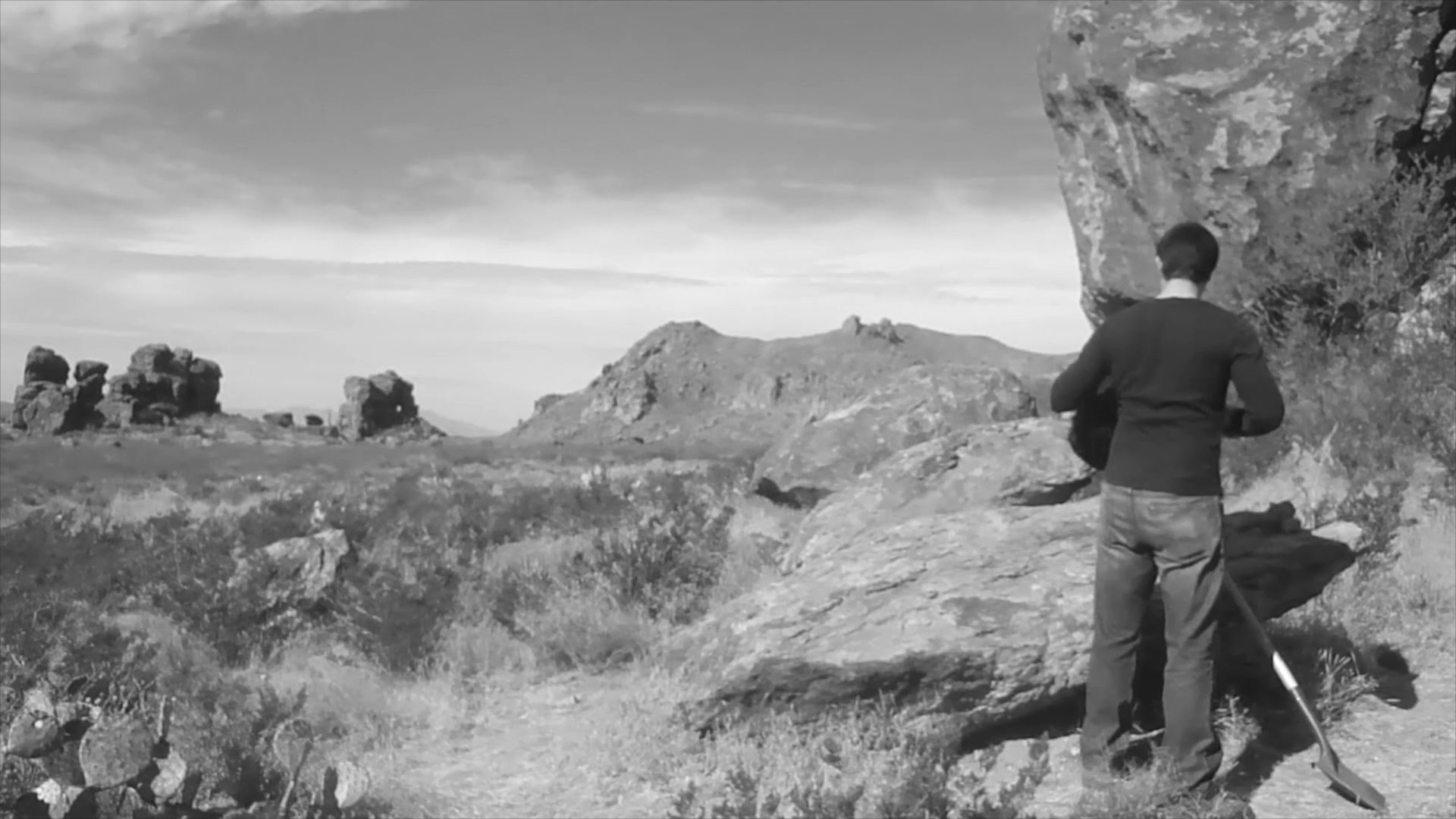 A researcher traverses a rugged, arid landscape carrying a shovel and backpack, eventually reaching a large rock formation to conduct an investigation.