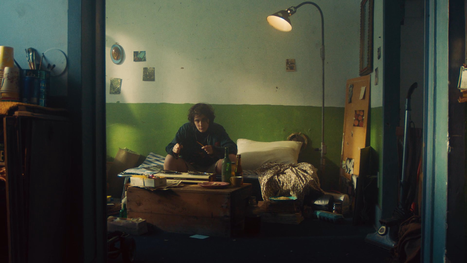 A young individual sits alone in a cluttered room, holding a pencil while lost in deep thought. The sequence shifts from a centered focus on the person to a closer profile view, highlighting a moment of quiet reflection.