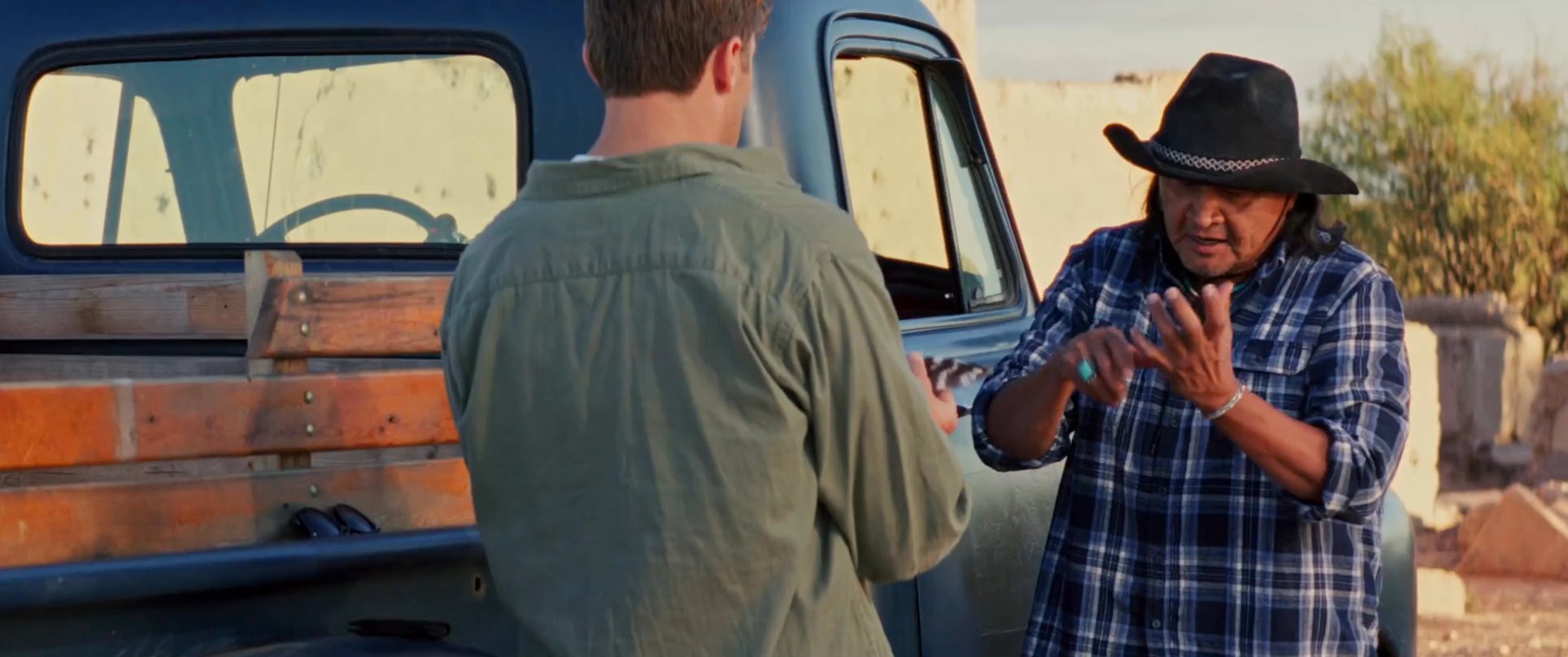 An Indigenous individual performs a traditional ritual, blessing a younger man with a feather beside a vintage pickup truck in an arid landscape. The interaction emphasizes cultural heritage, guidance, and a solemn, respectful exchange.