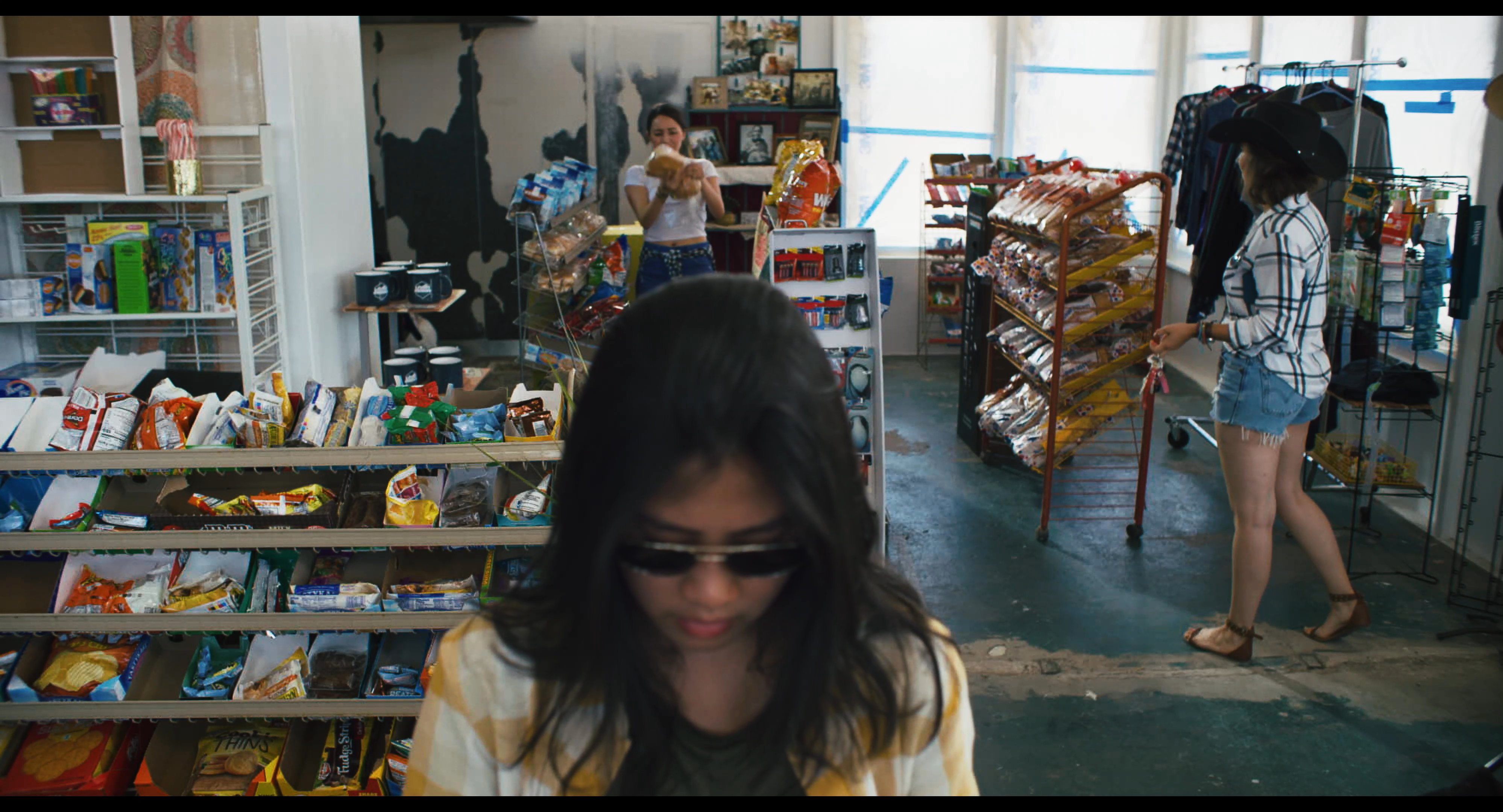 A woman enters a rural gas station convenience store while a vehicle remains parked outside. The interior scene captures a casual shopping moment as she adjusts her sunglasses and looks around the store shelves.