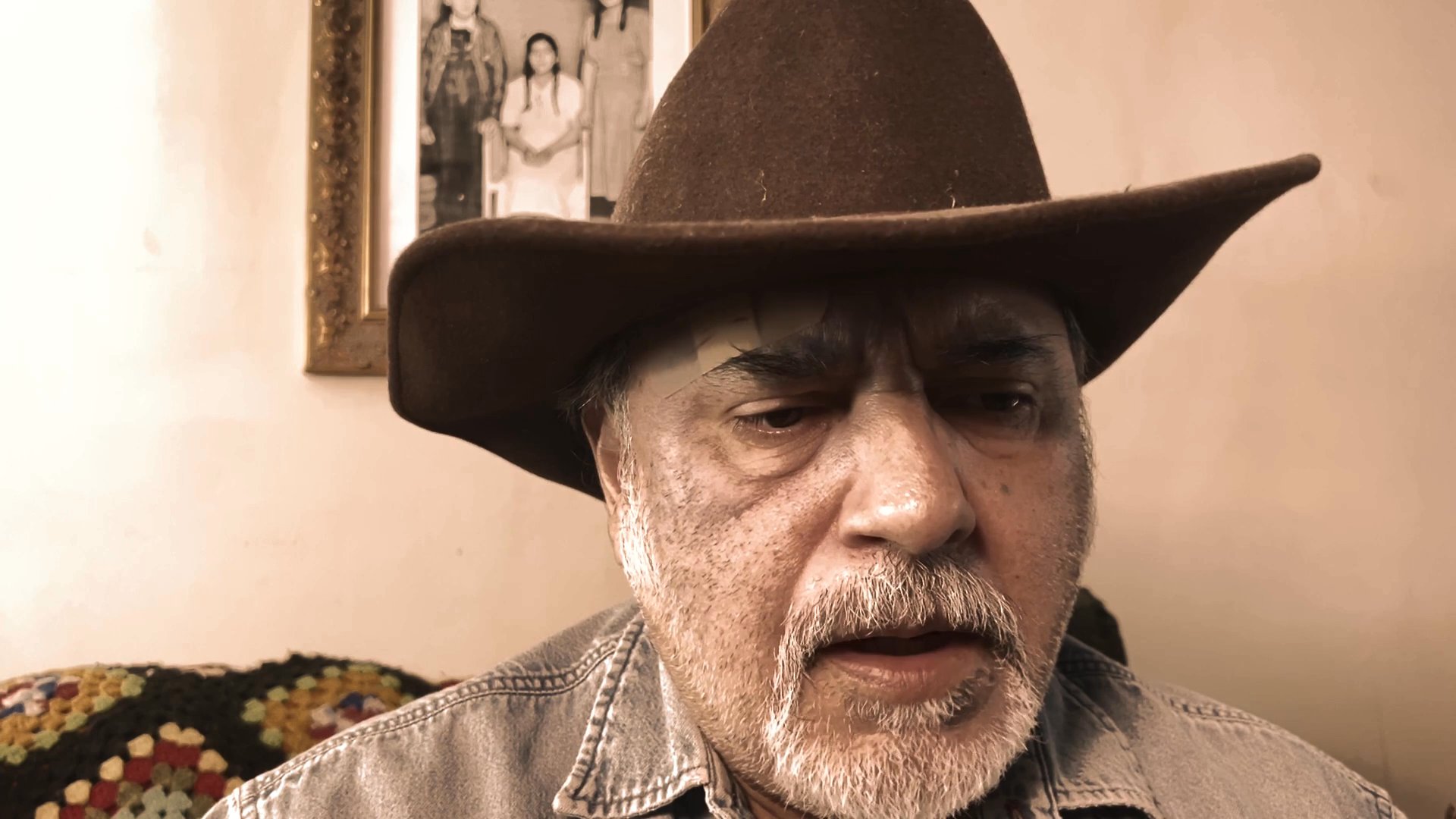An older man wearing a cowboy hat delivers a focused monologue, his facial expressions shifting as he communicates to an off-camera audience.