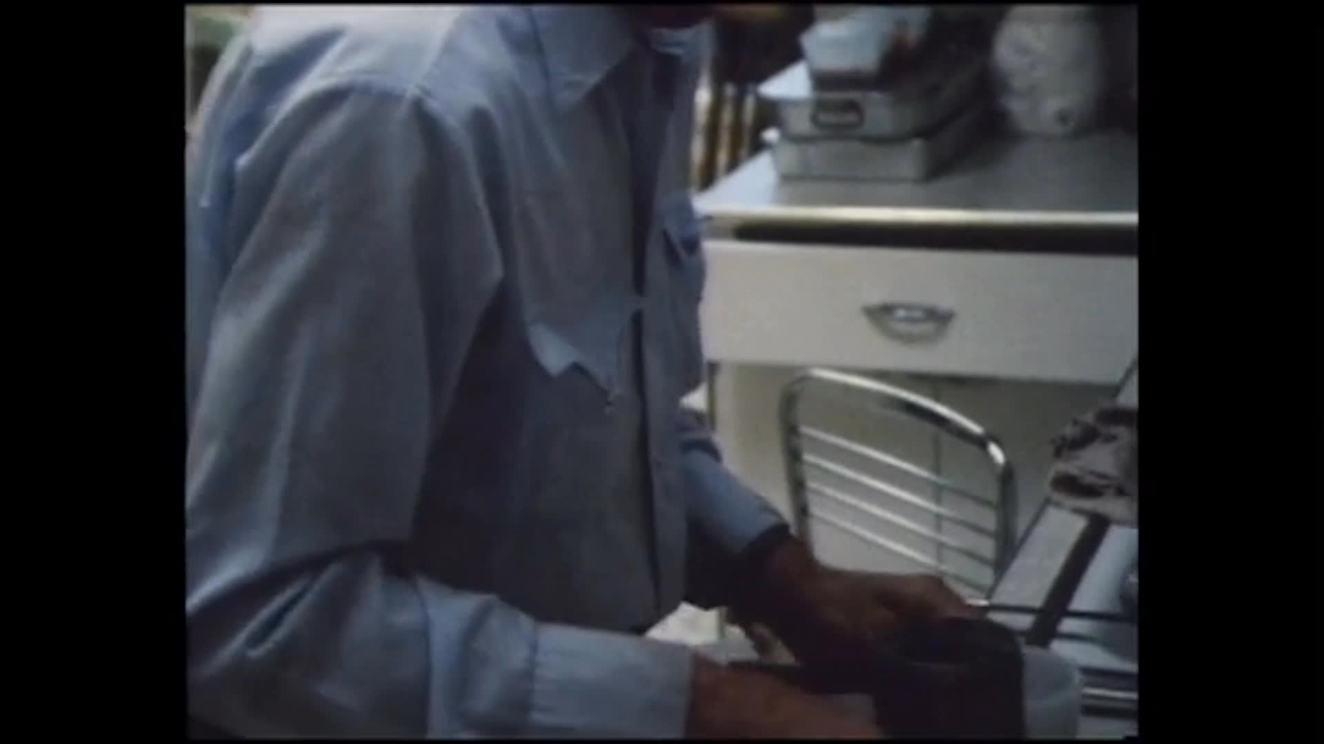 A man prepares breakfast in a domestic kitchen, tending meat in a frying pan and handling food at the counter. He finishes by carrying a tray of prepared portions.