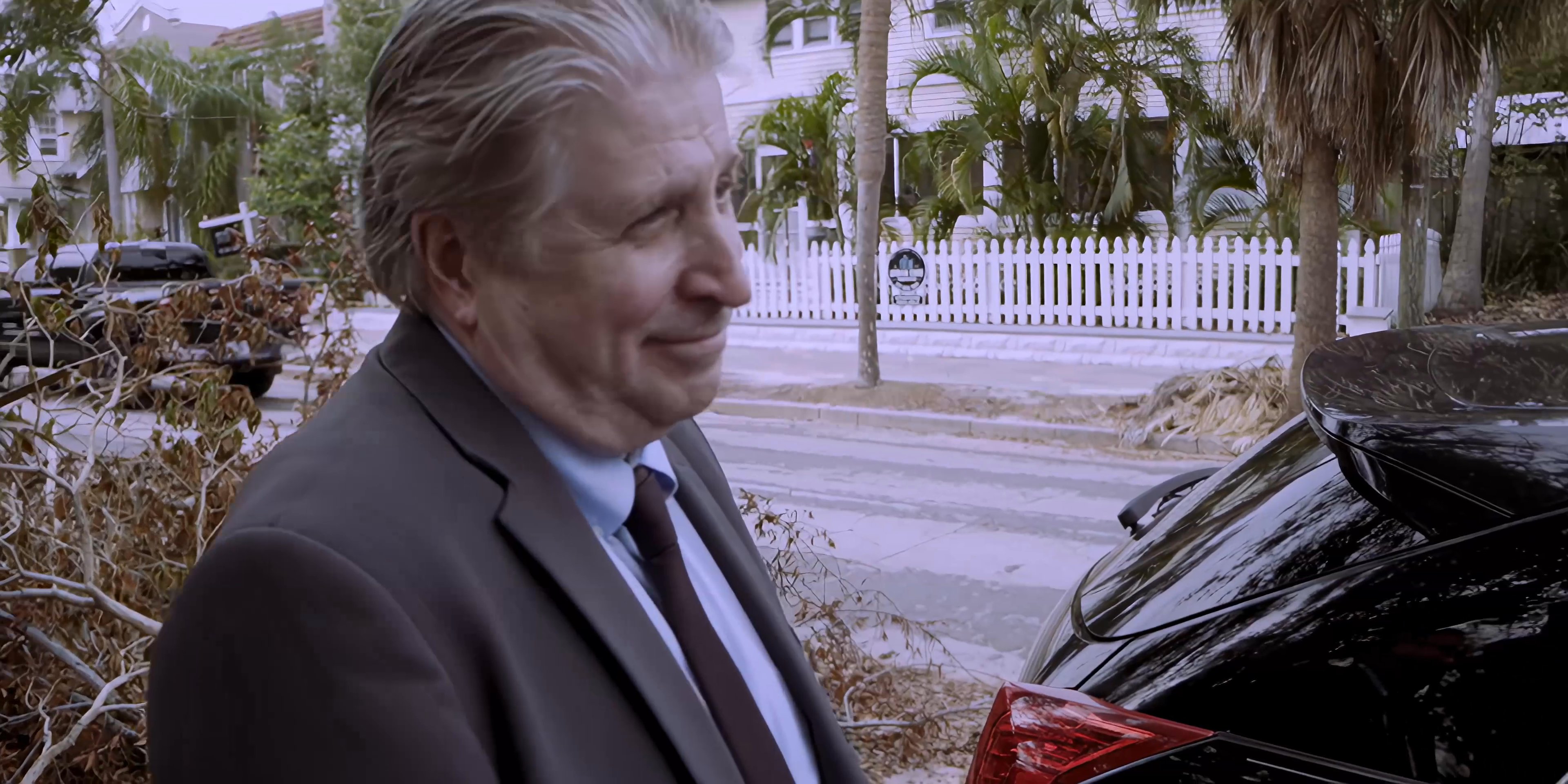 An older man in a suit approaches a younger man wearing a casual patterned shirt on a residential street. They engage in a brief conversation amidst the backdrop of a quiet neighborhood.