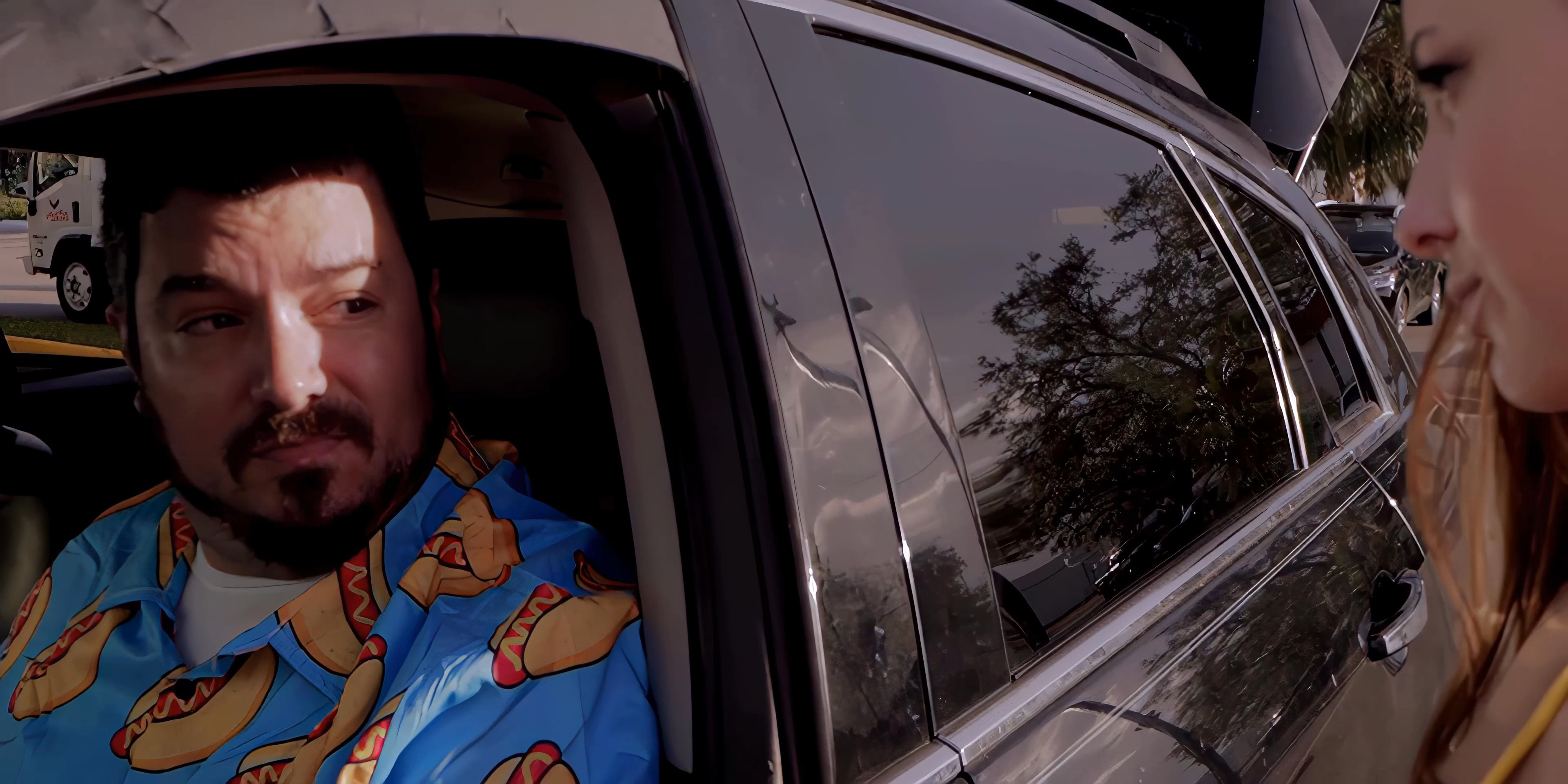A woman engages in a conversation with a man sitting in the driver's seat of a car, while an older man in a suit looks on nearby in an outdoor parking lot.