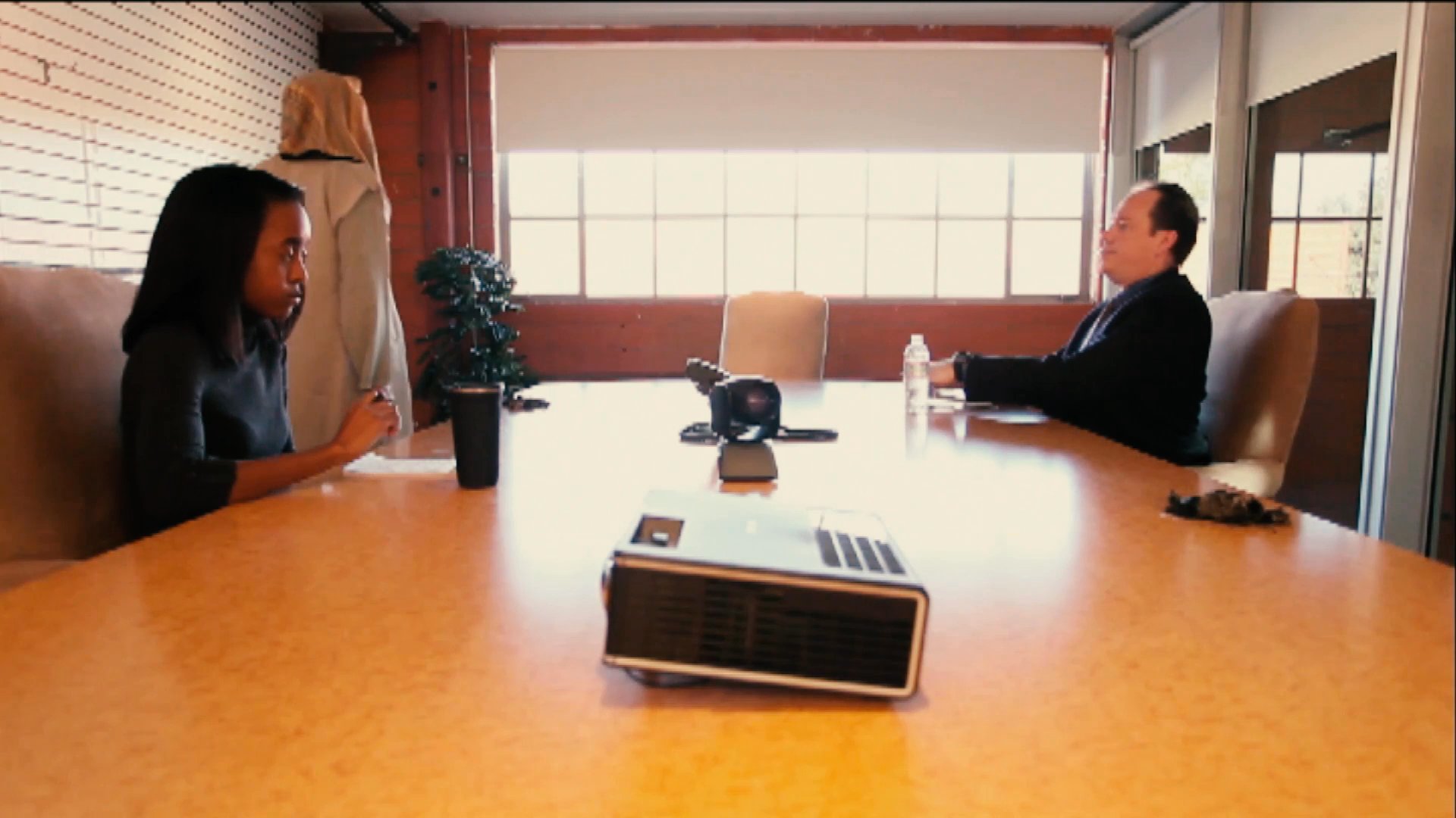 Two professionals engage in a formal meeting within a conference room setting, interrupted by a figure walking through the background.
