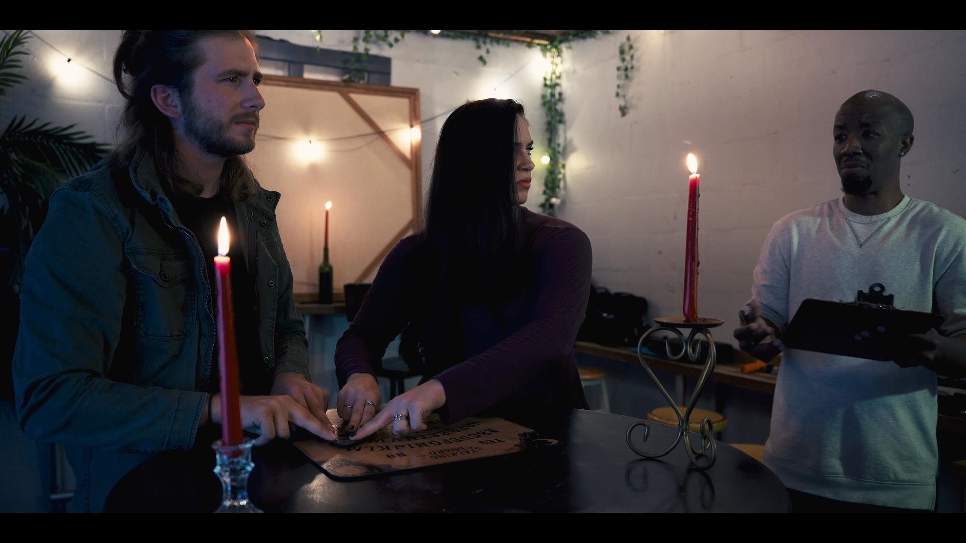 Three individuals participate in a seance around a candlelit table, focusing intently on a ouija board while one person monitors the activity with a clipboard. The atmospheric, dimly lit setting builds a sense of mystery and paranormal intrigue.