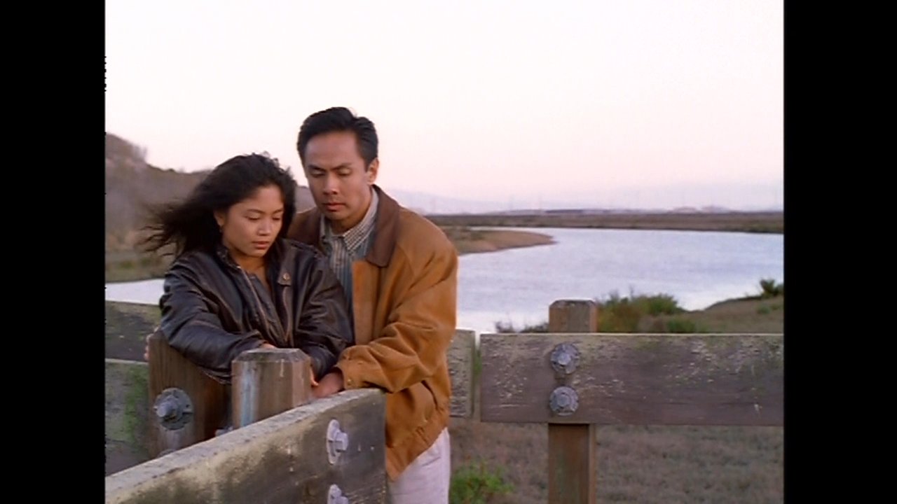 A couple stands in an intimate embrace beside a waterfront marshland during sunset. The pair shares a quiet moment together as the sky turns hues of dusk over the distant horizon.