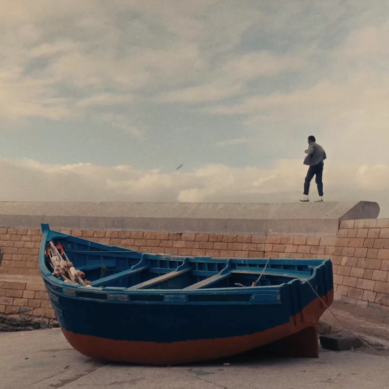 Video of man walking along cliff wall next to the sea with wooden blue boat in the foreground