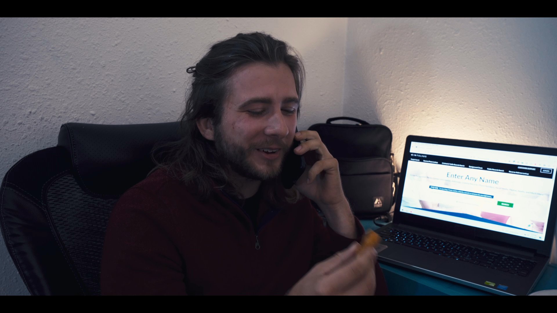 A man manages a professional phone conversation while snacking and working in a casual home environment. The scene alternates between his workspace and a dimly lit bedroom setting.