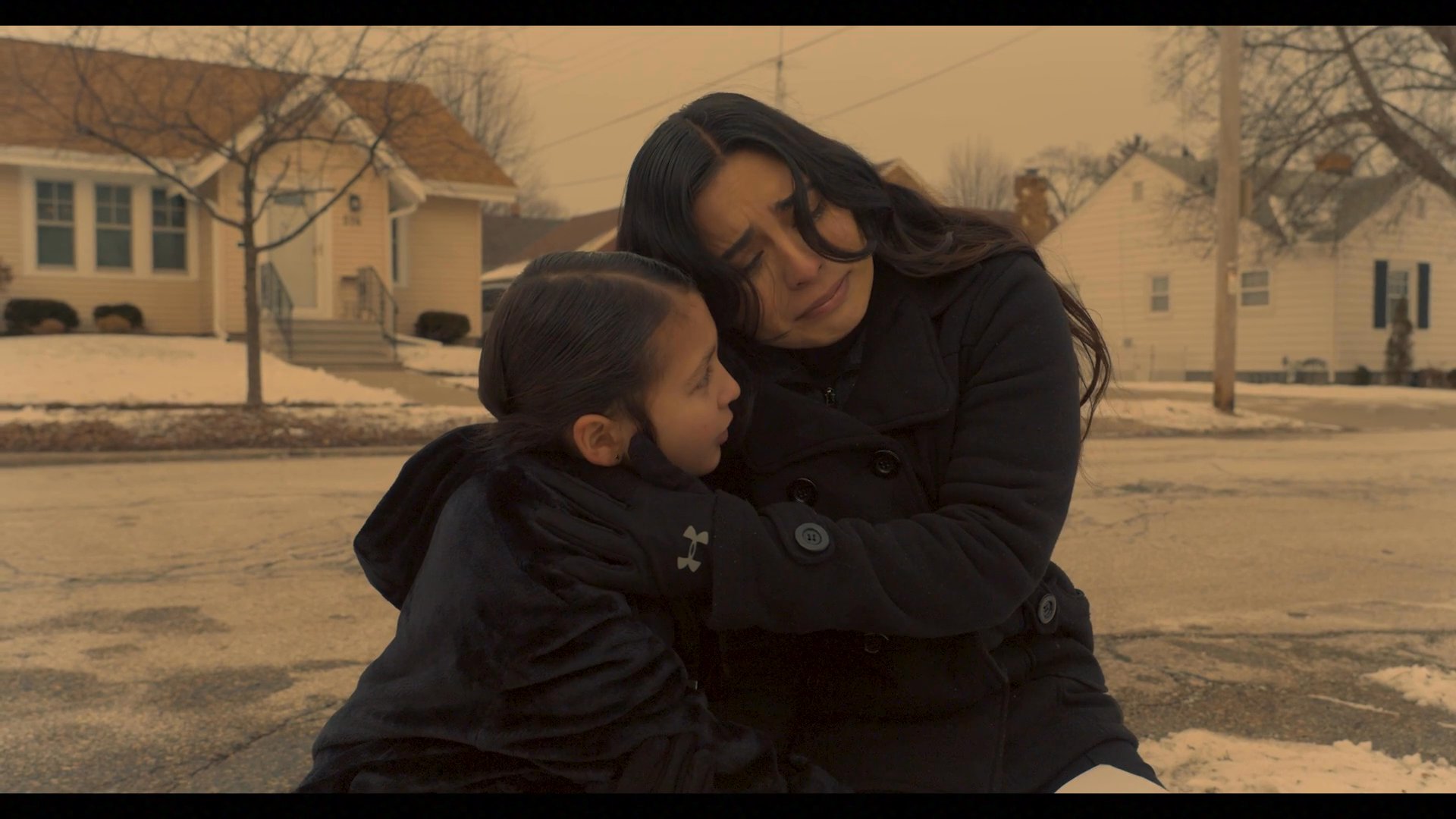 Two individuals console one another while sitting before a roadside memorial in a winter suburban setting. The scene captures a profound moment of shared grief and mutual emotional support.