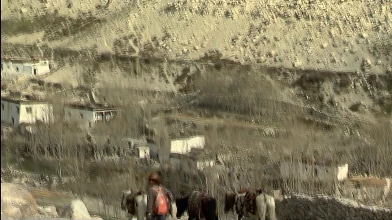 Residents of a remote Himalayan village gather near a monastery as a local man reflects on regional life and traditions. The scenery highlights the stark, arid mountain terrain characteristic of the Mustang region.