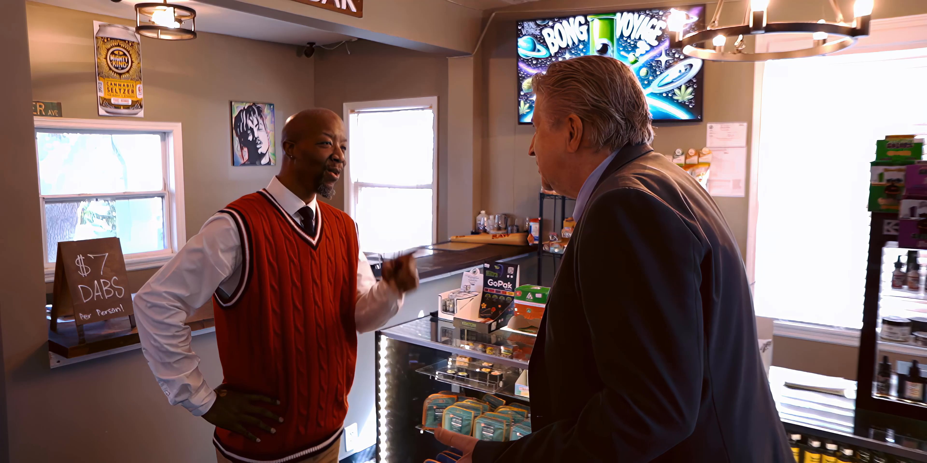 A shopkeeper in a red sweater greets and consults with a customer in a suit behind a retail service counter. The two engage in a professional conversation within a well-lit commercial space.