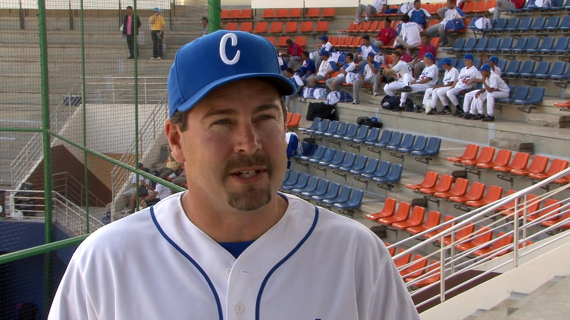 A baseball coach wearing a team uniform discusses professional training in a stadium setting as players practice on the field behind him.