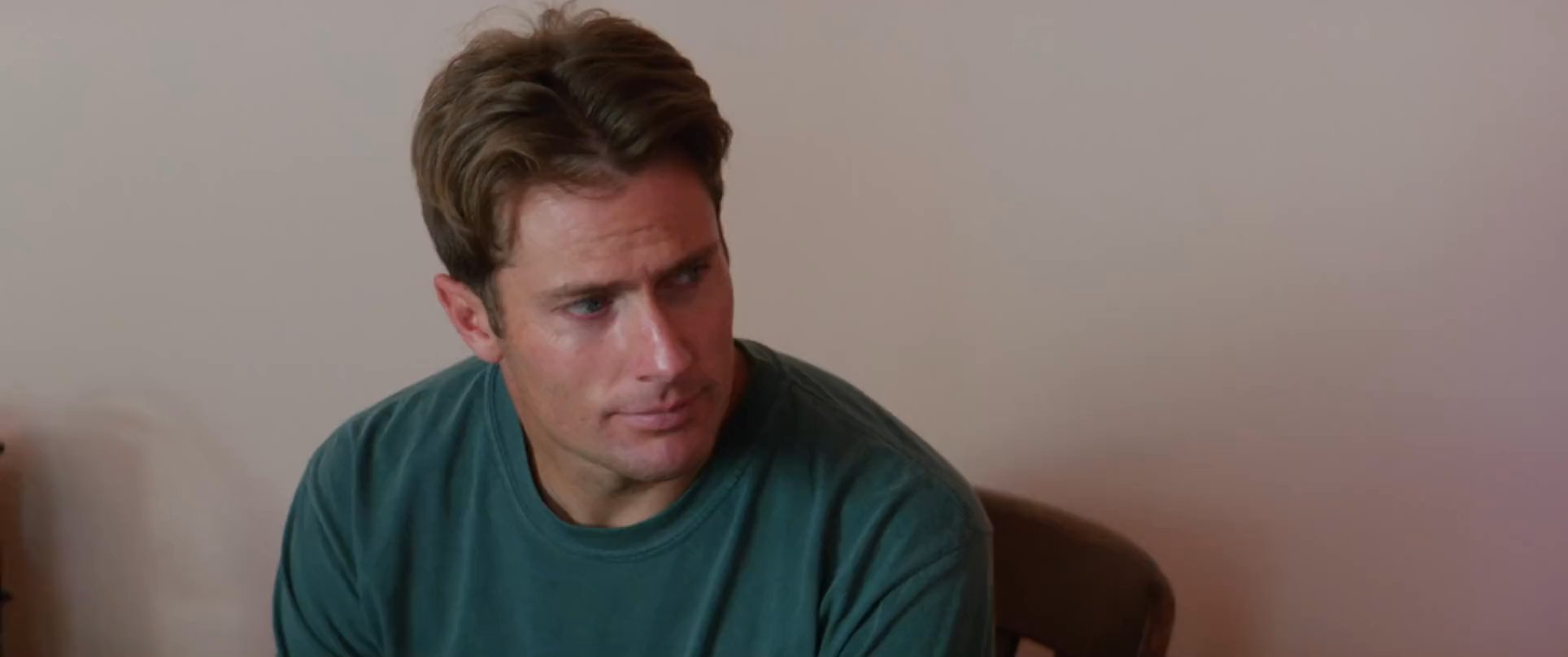 Two men have a somber conversation in a bedroom while a vintage television displays controversial messaging in the background. The scene captures a tense moment of interpersonal reflection amidst external societal commentary.
