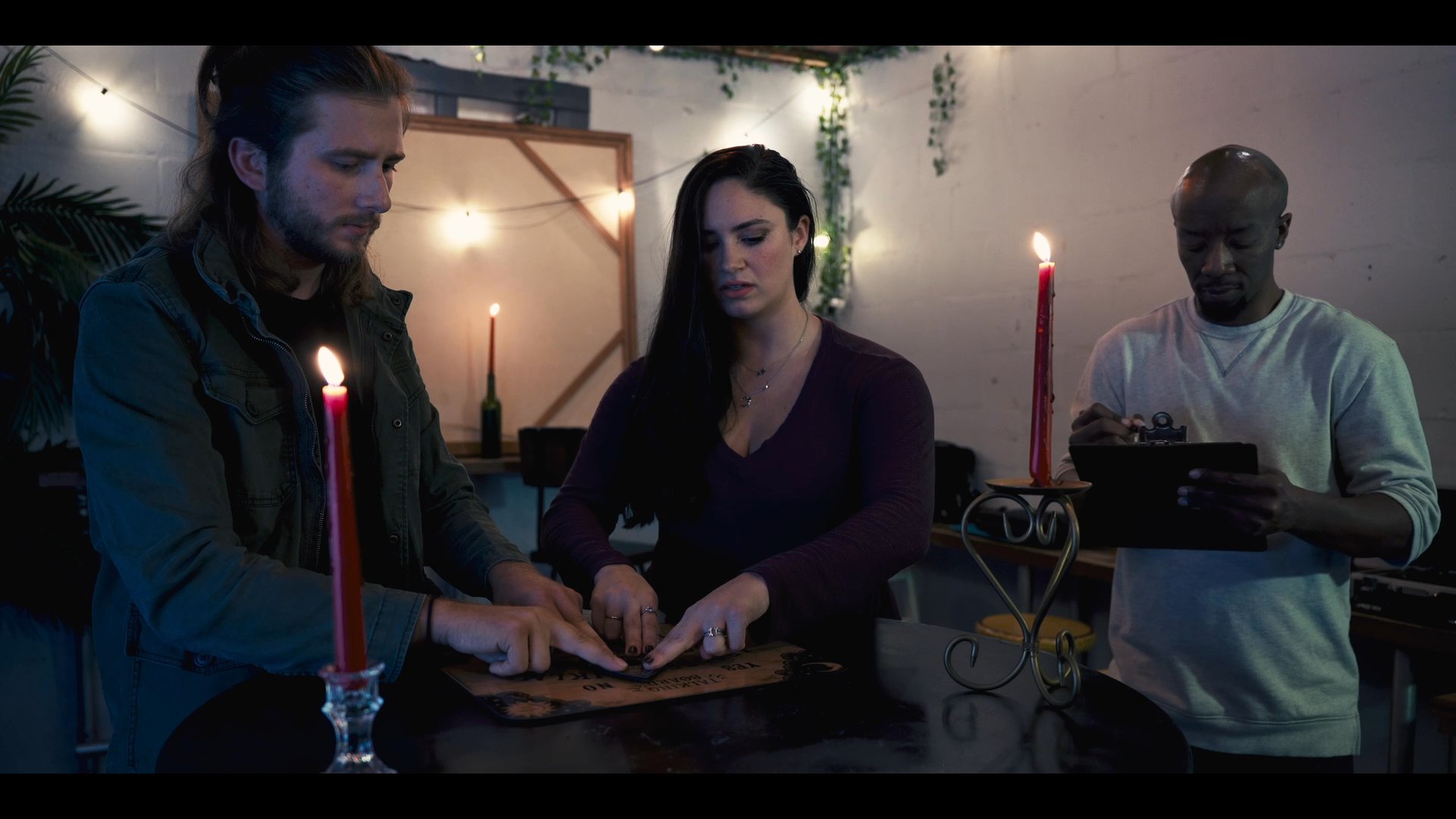 Three individuals participate in a seance, using a ouija board by candlelight to facilitate potential spirit communication. A fourth person observes and documents the interaction, maintaining a tense and focused atmosphere.