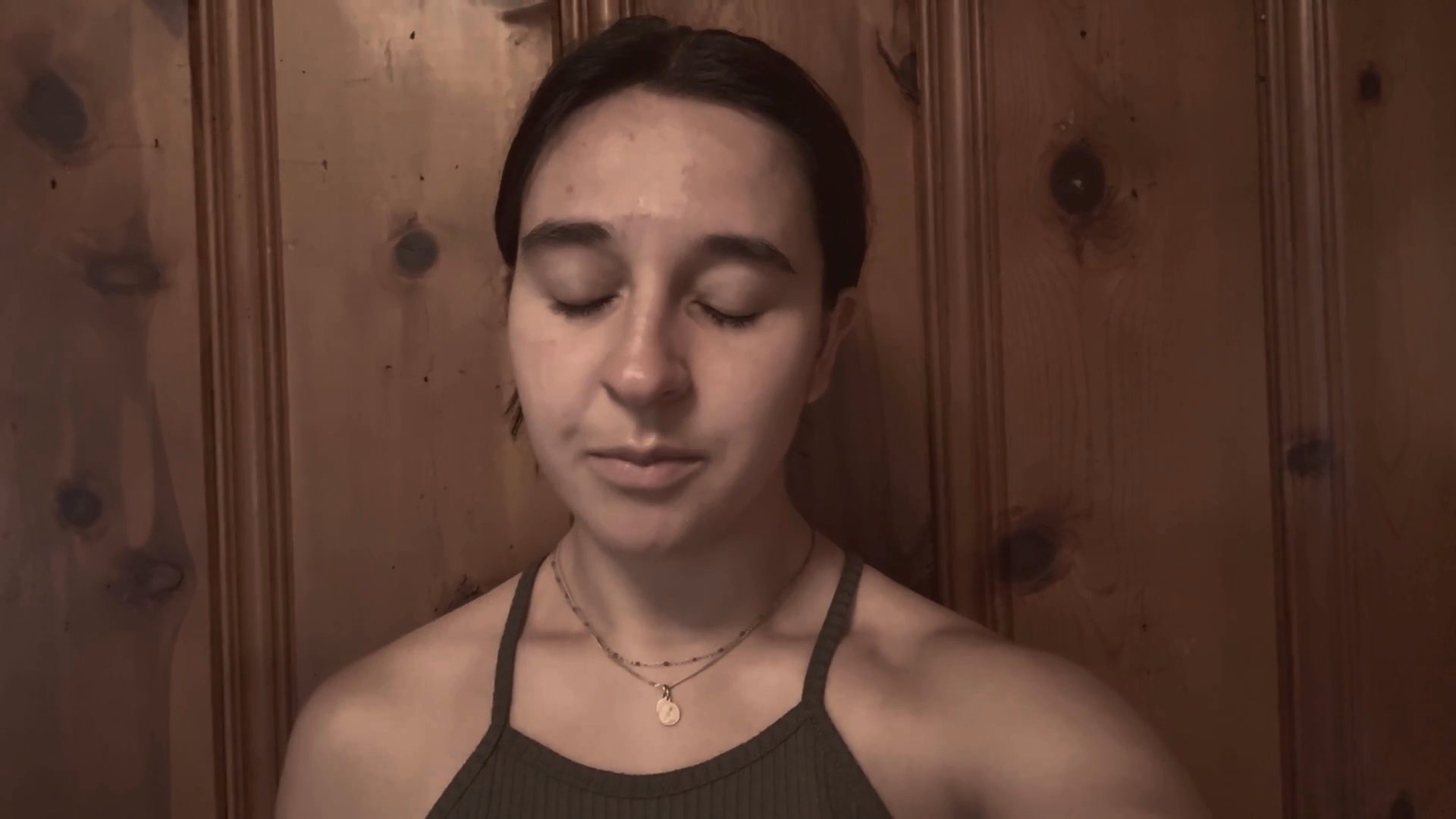A woman speaks directly to the camera while positioned in front of a wood-paneled wall, delivering a conversational monologue.