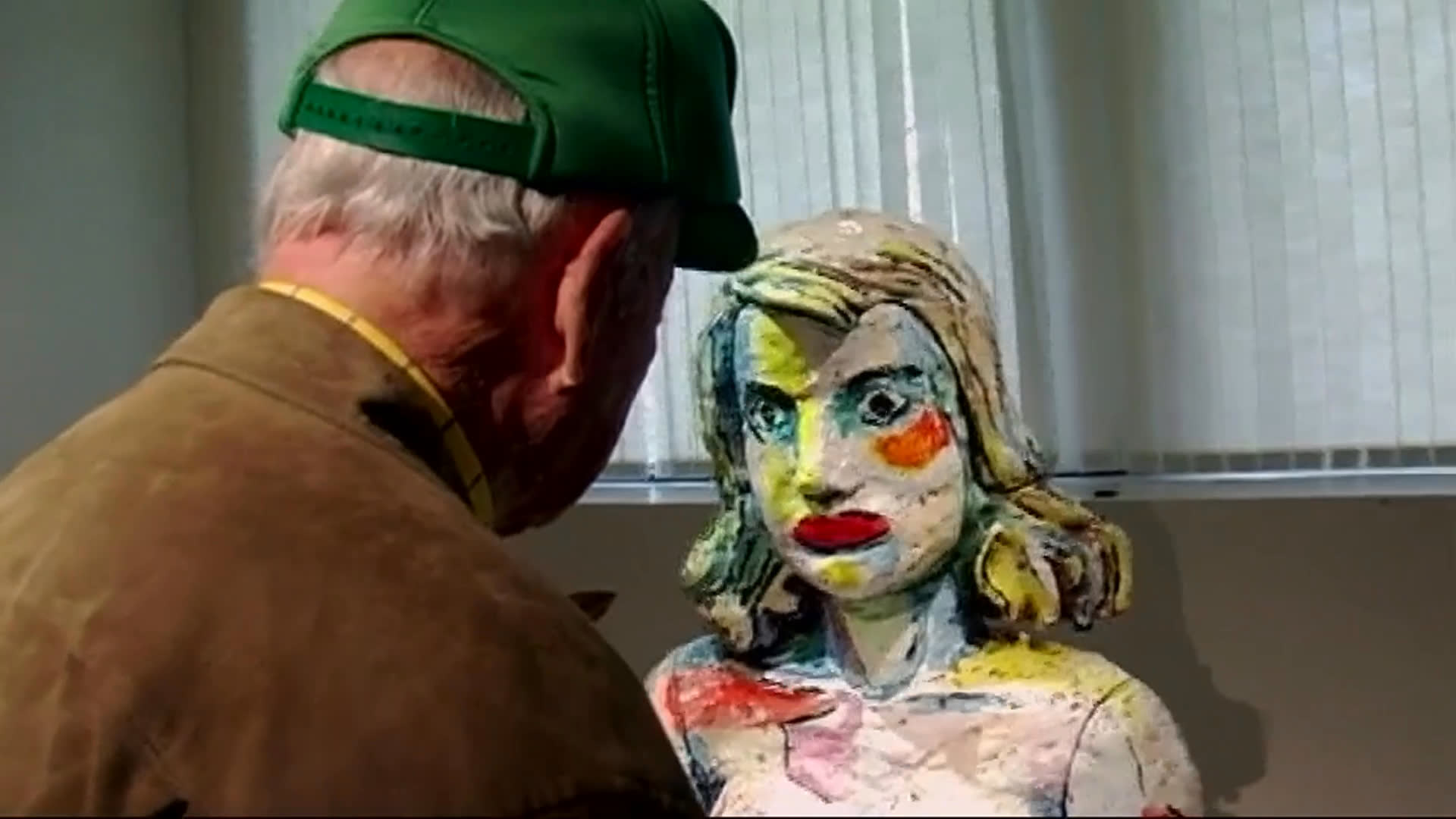 An elderly man in a green cap examines a vividly painted seated figure sculpture in a gallery, accompanied by a woman. He moves around the artwork and walks farther into the museum as the sculpture remains in the foreground.