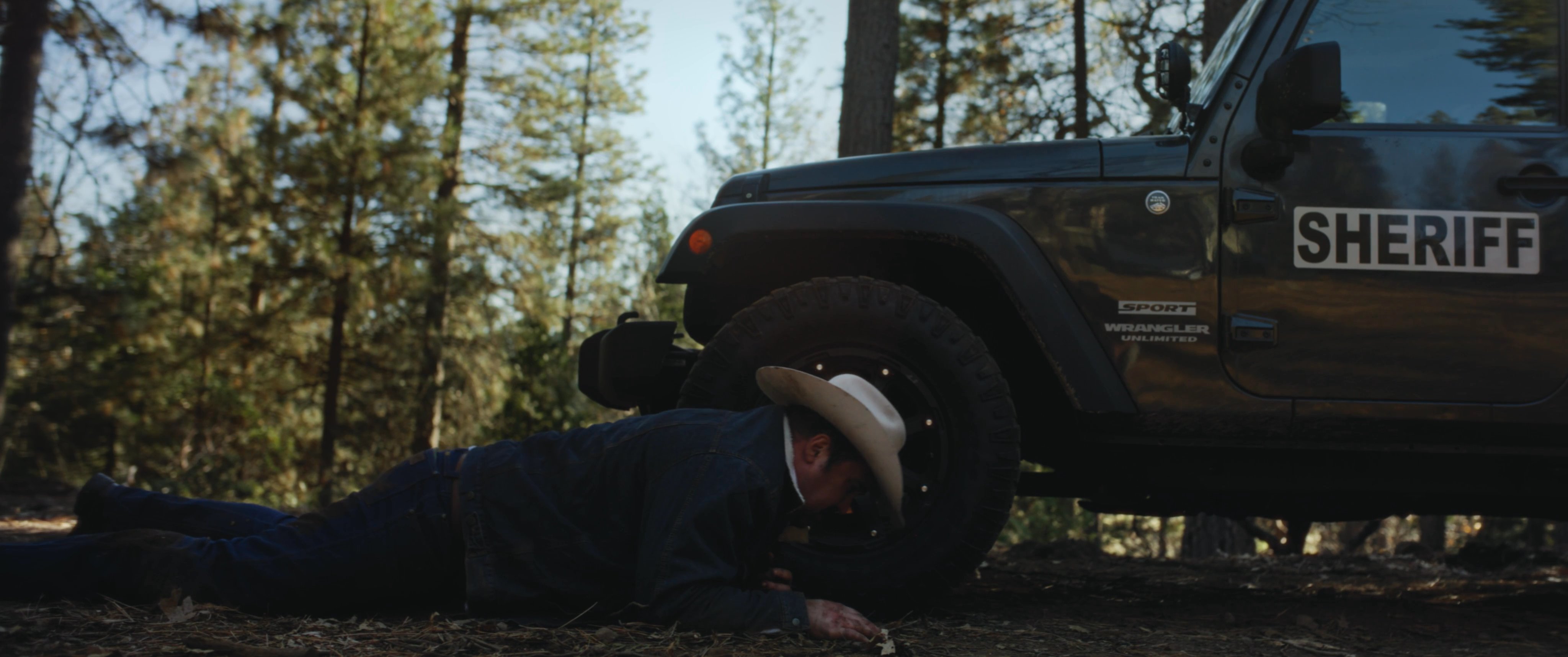 An injured man wearing a cowboy hat struggles to crawl across the ground toward a parked sheriff vehicle in a wooded environment.