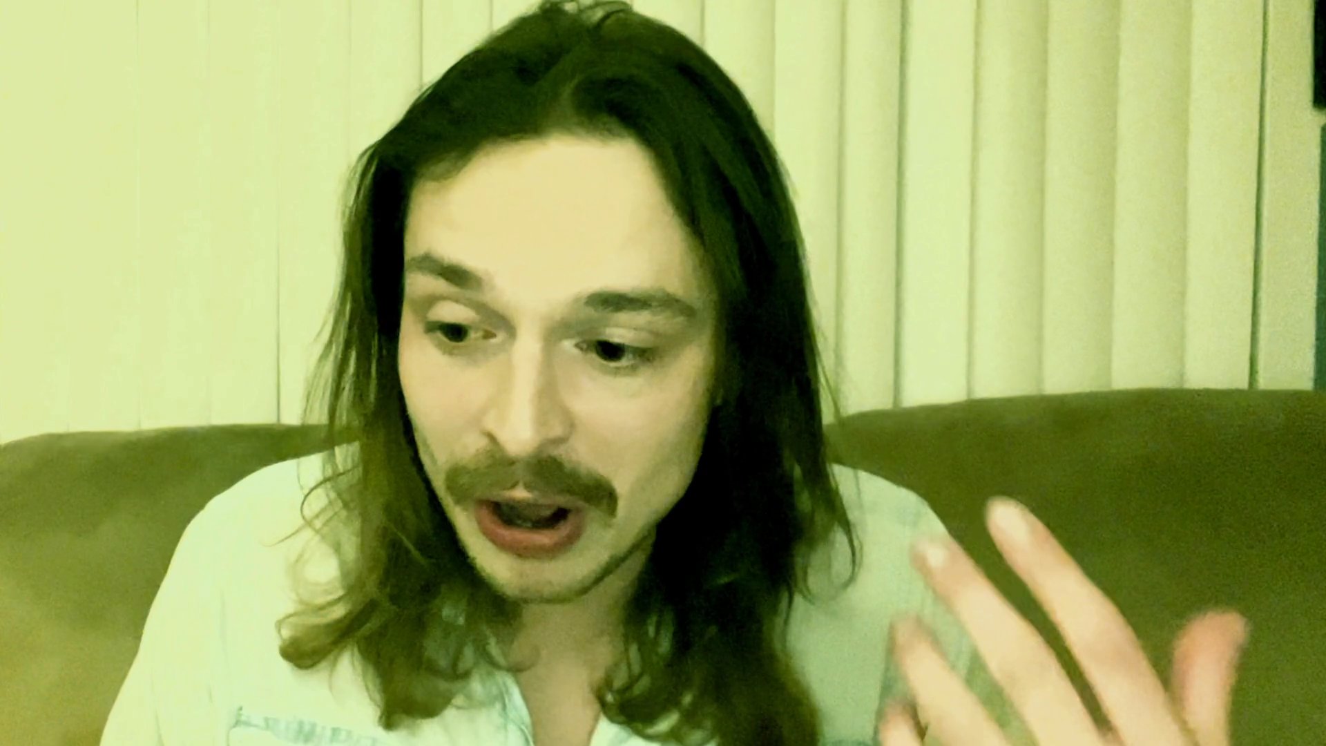 A man with long hair and a mustache engages in an animated, expressive monologue while seated indoors. He uses hand gestures and varied facial expressions to convey his narrative to an off-camera audience.