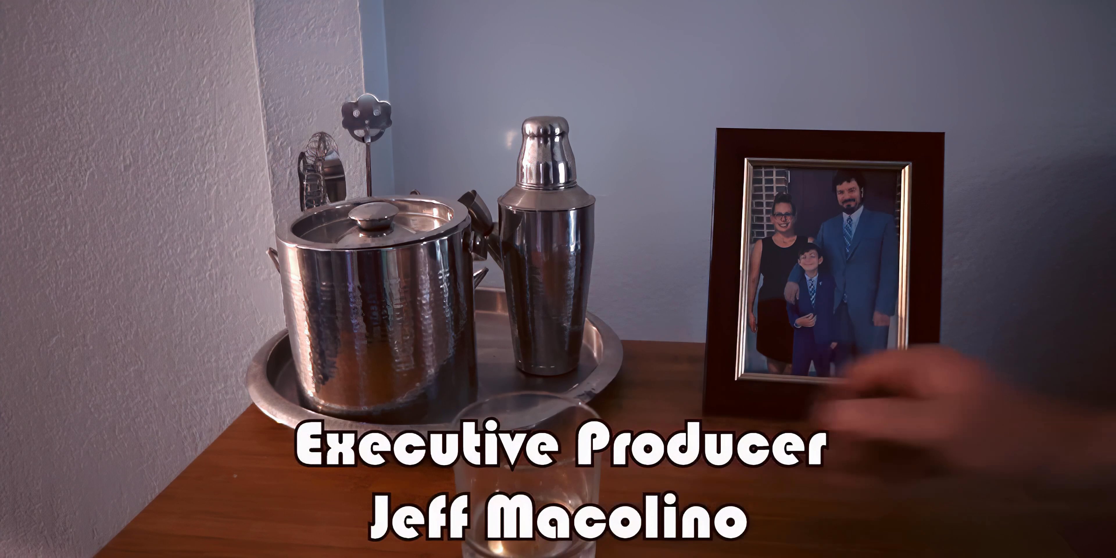 A man in a home environment walks through a living room and interacts with a framed photograph near a bar setup during the opening title sequence.