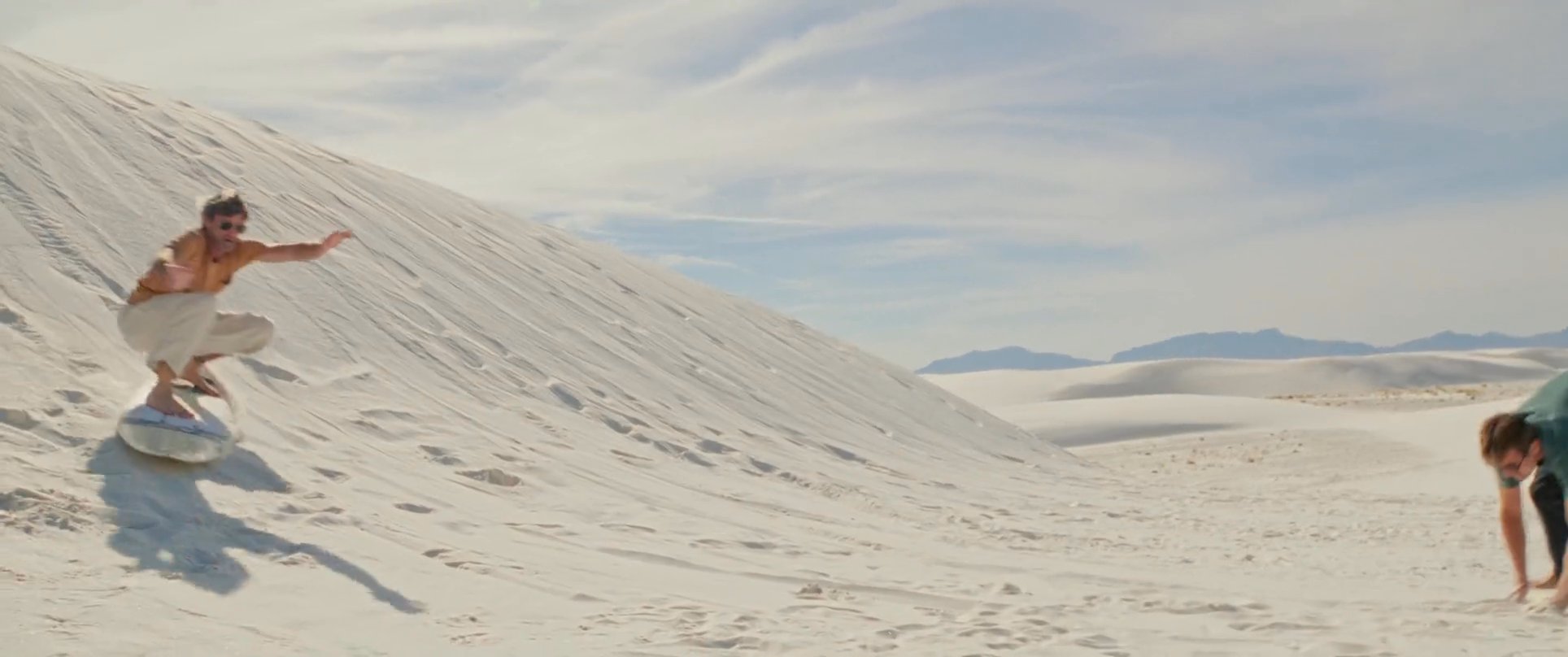 Athletes navigate the steep slopes of white gypsum dunes on sandboards in a vast desert landscape. The sequence captures high-speed descents followed by celebratory moments of successful runs.