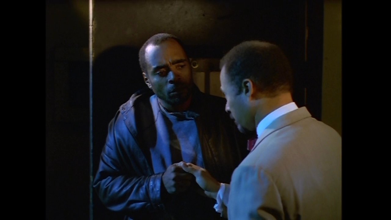 Two men engage in a serious, quiet conversation in a dimly lit office environment. They exchange information and gestures as they coordinate their next steps.