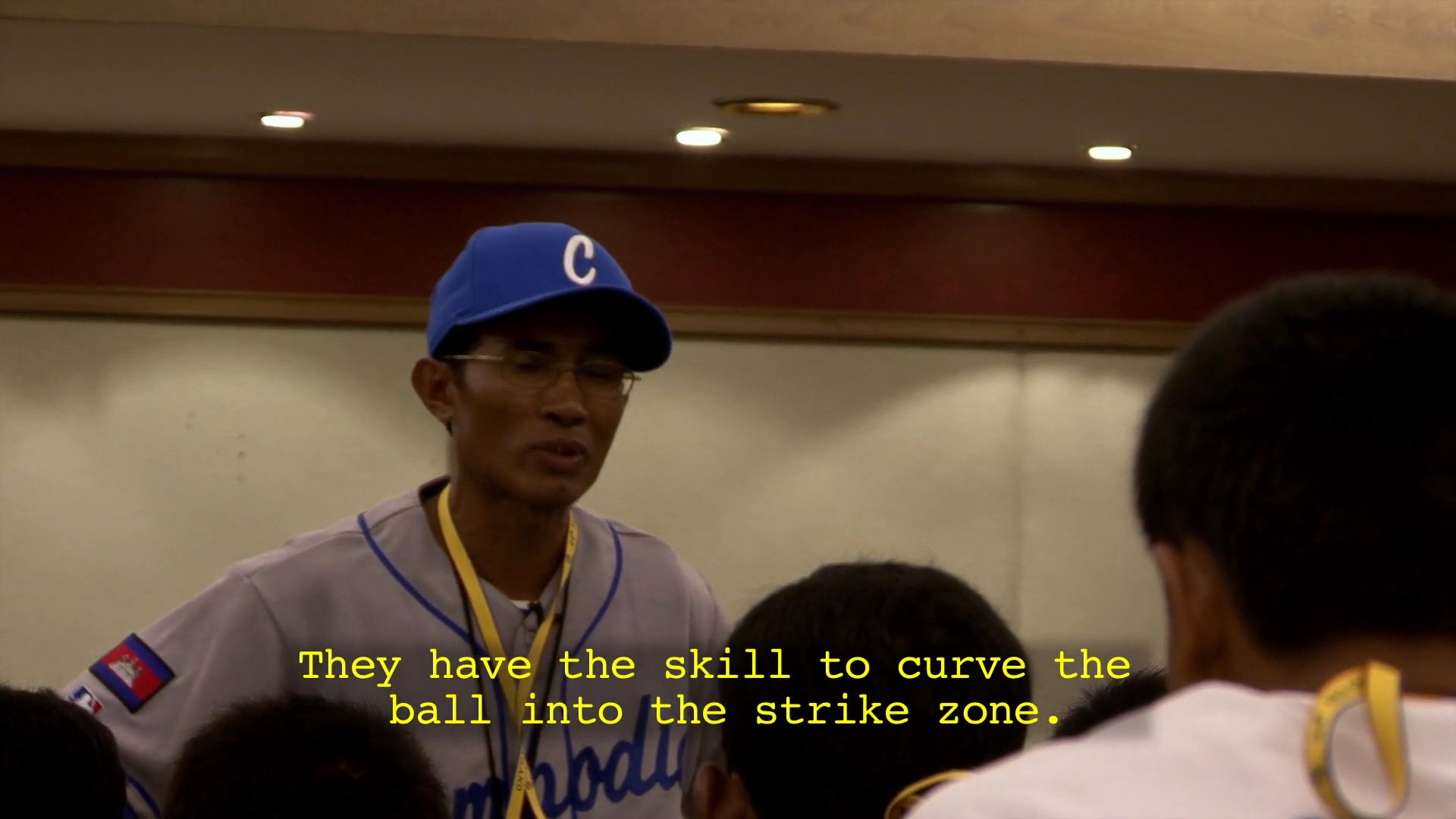 A coach wearing a baseball uniform addresses his team in a conference room, providing tactical instruction on handling specific pitches during gameplay.