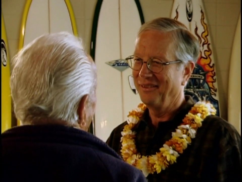Two individuals engage in a warm, friendly conversation while others attend a social event in an indoor setting decorated with surfboards.