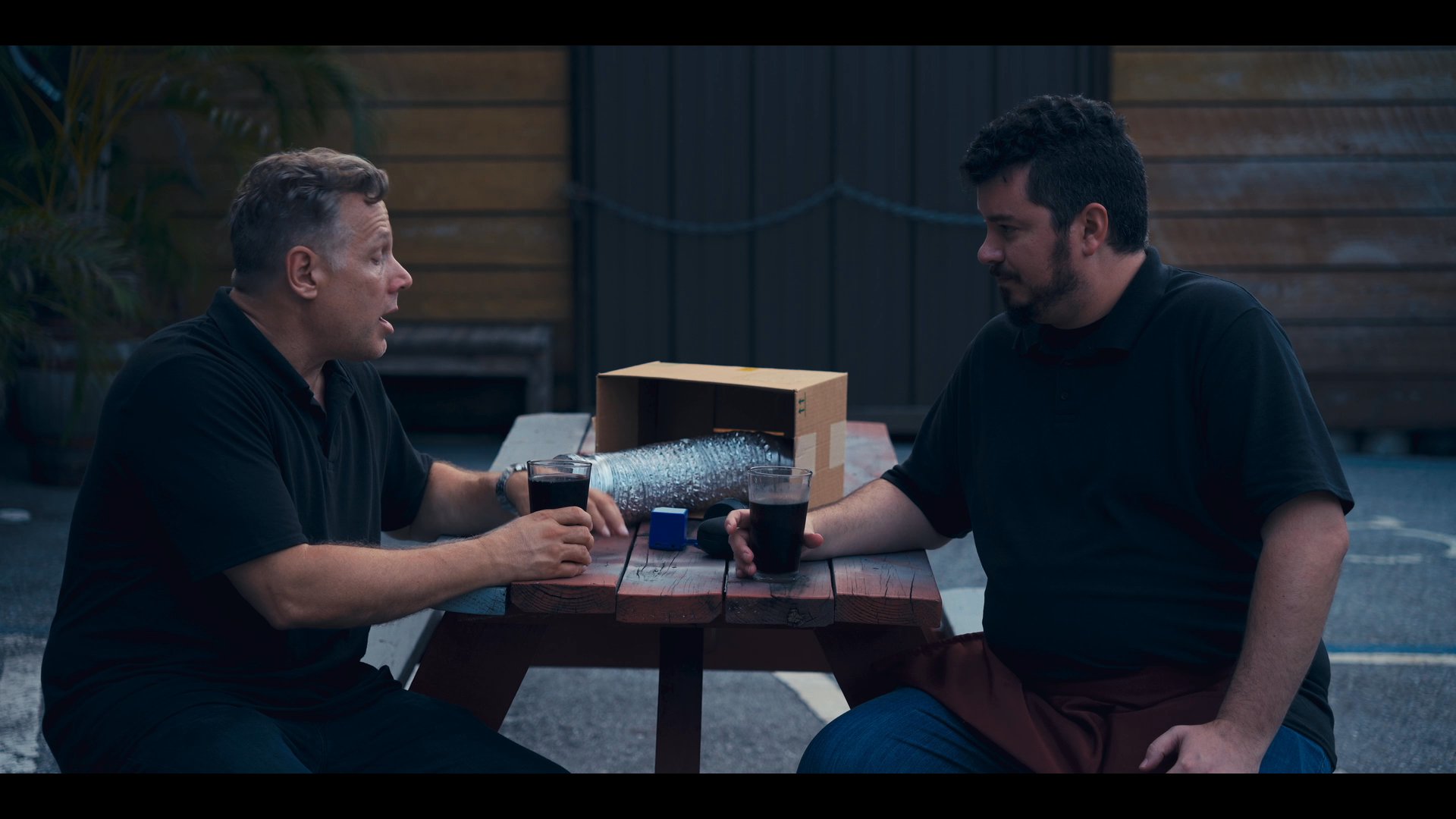 Two men sit at an outdoor table engaged in a focused conversation while sharing beverages.